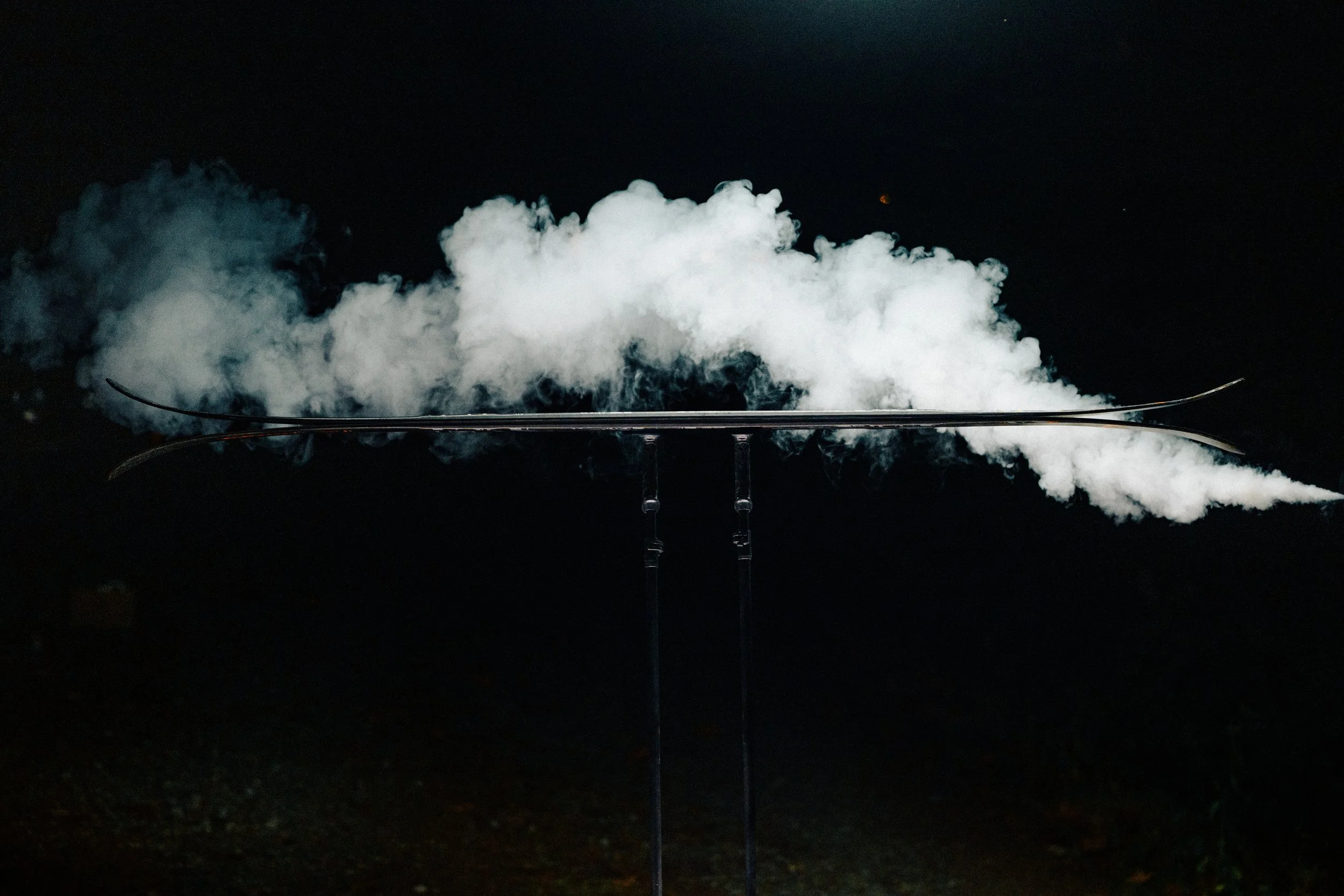 Nighttime scene with a trail of white smoke or vapor against a dark background, with ski or snowboard equipment visible at the bottom.