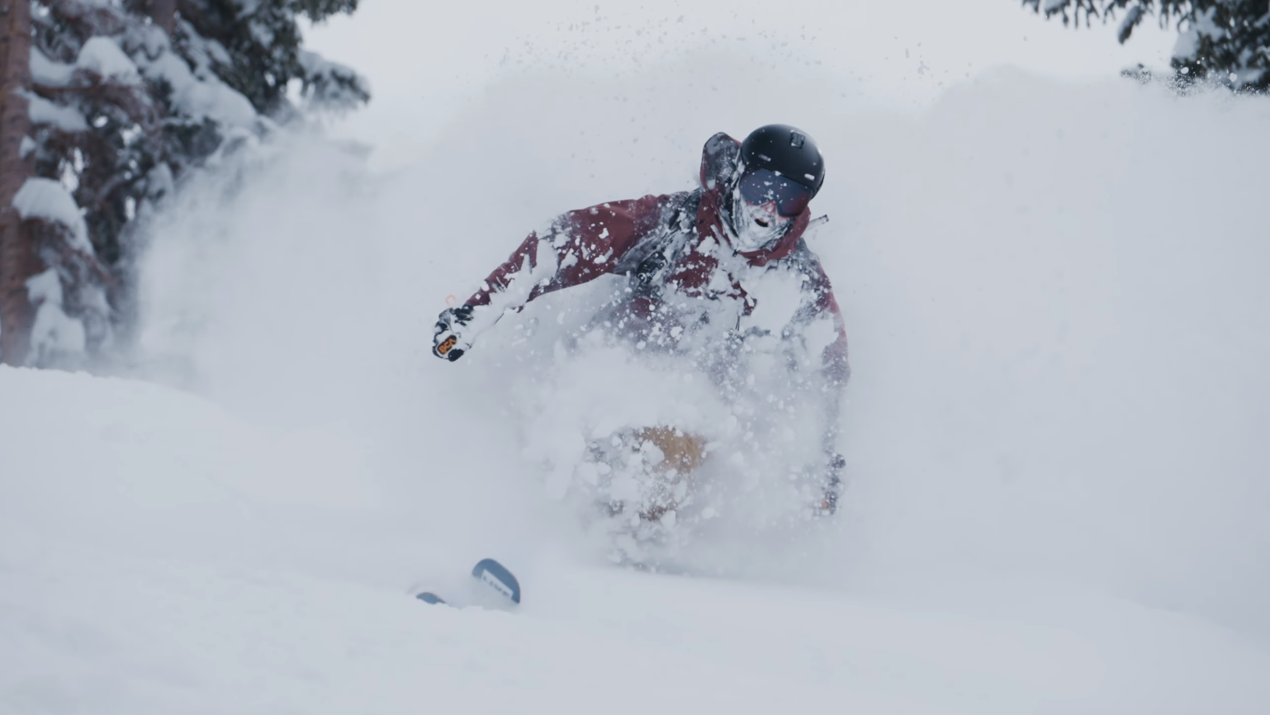 A person wearing a helmet and winter clothing snowboarding or skiing downhill in deep snow amidst snowy trees. A person getting a deep powder turn.