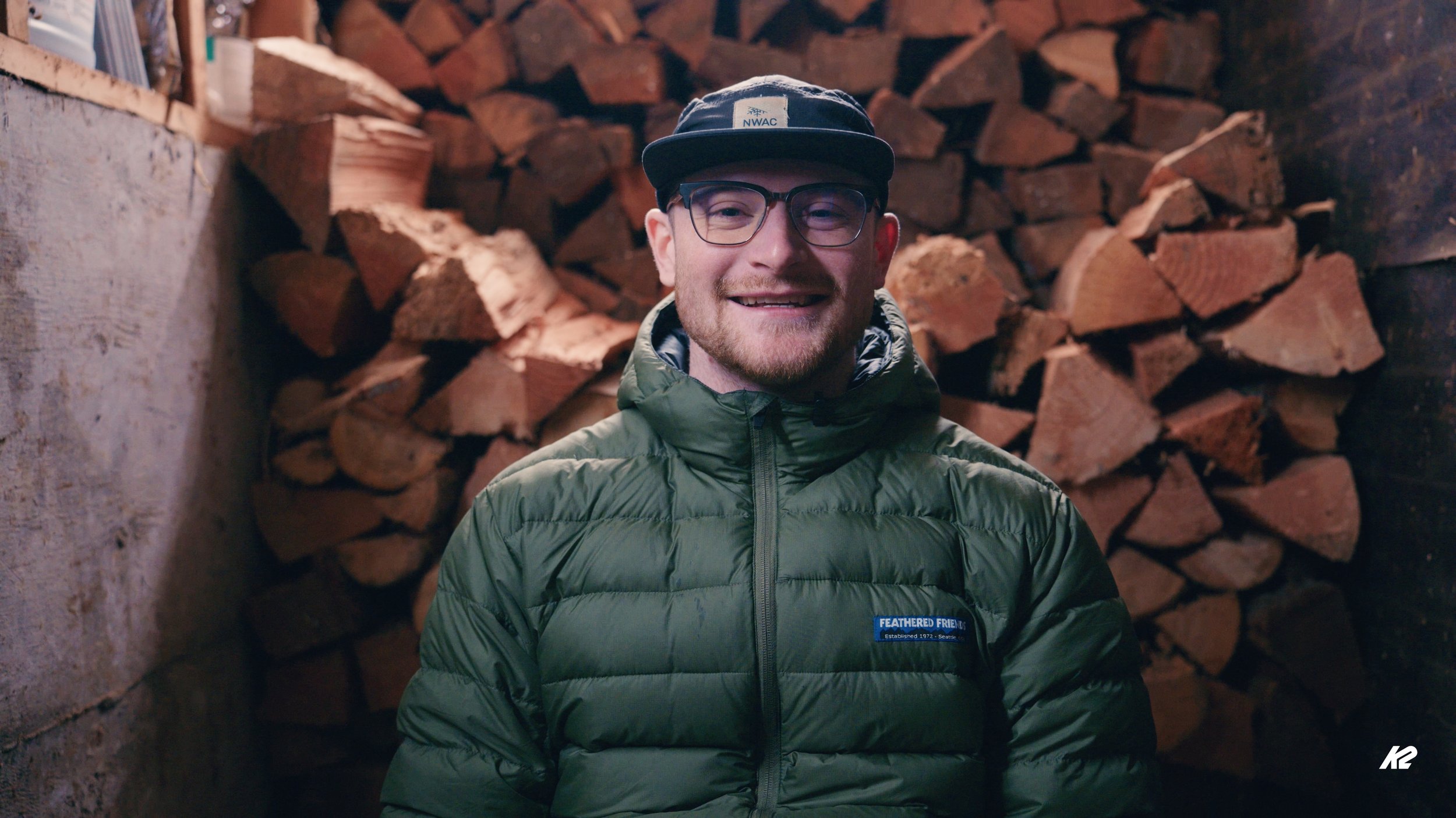 A man wearing glasses, a black cap, and a green puffy jacket standing inside a wood storage area with stacked firewood in the background.