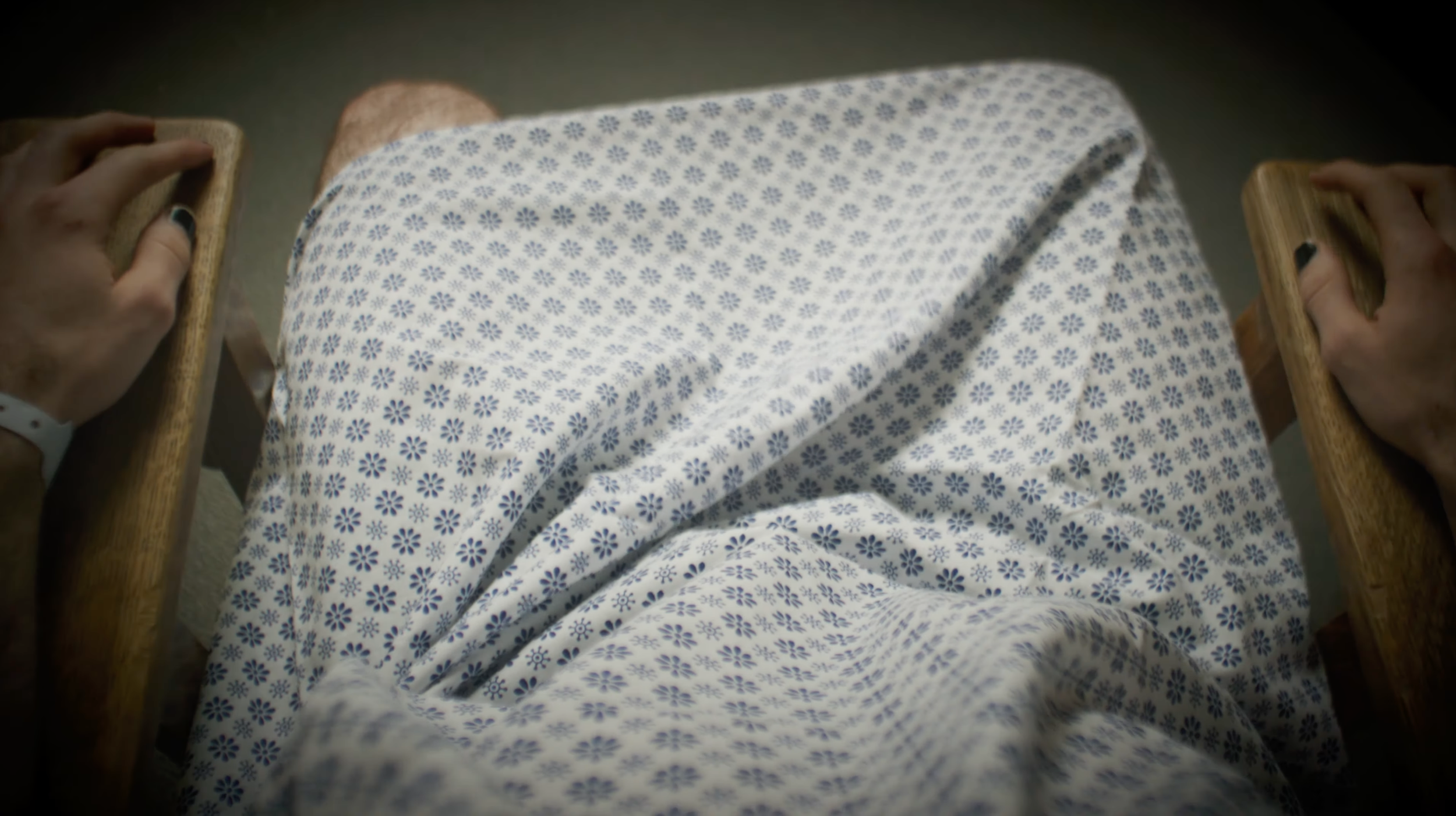 Person lying in a hospital bed wearing blue and white floral pajamas, holding wooden side rails of the bed.
