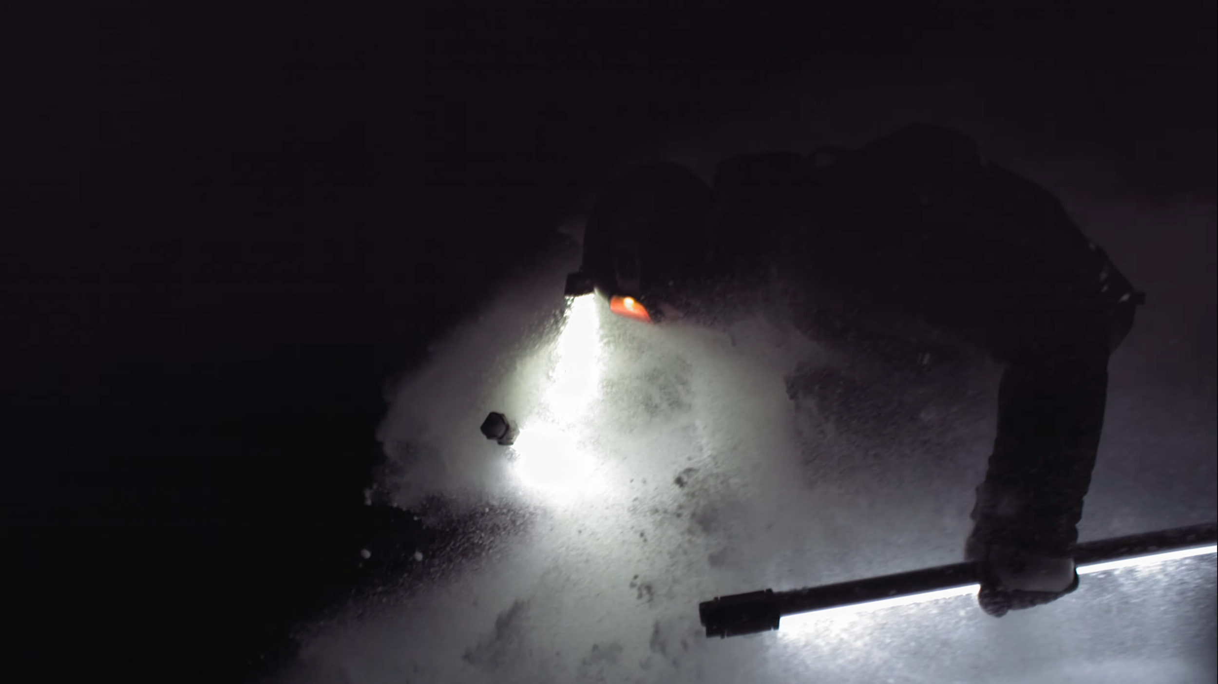 A person shoveling snow at night, illuminated by a bright light, creating a stark contrast against the dark background.