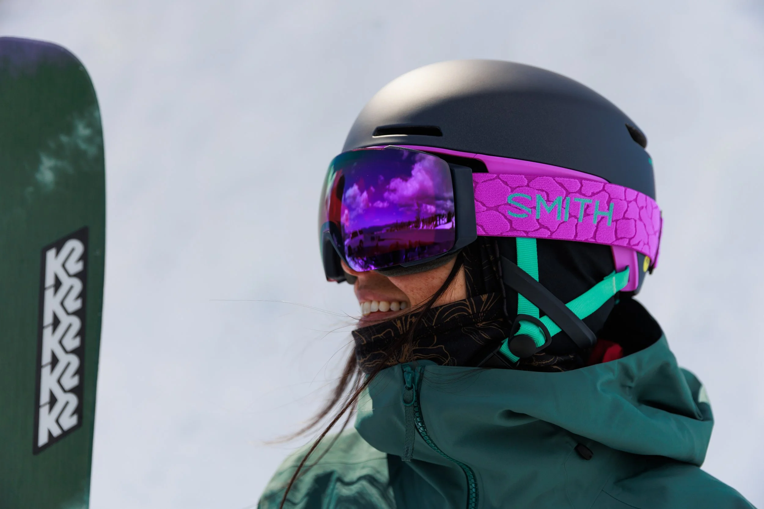A person smiling while wearing ski gear, including a black helmet, purple and teal goggles, and a teal jacket, on snowy slopes.