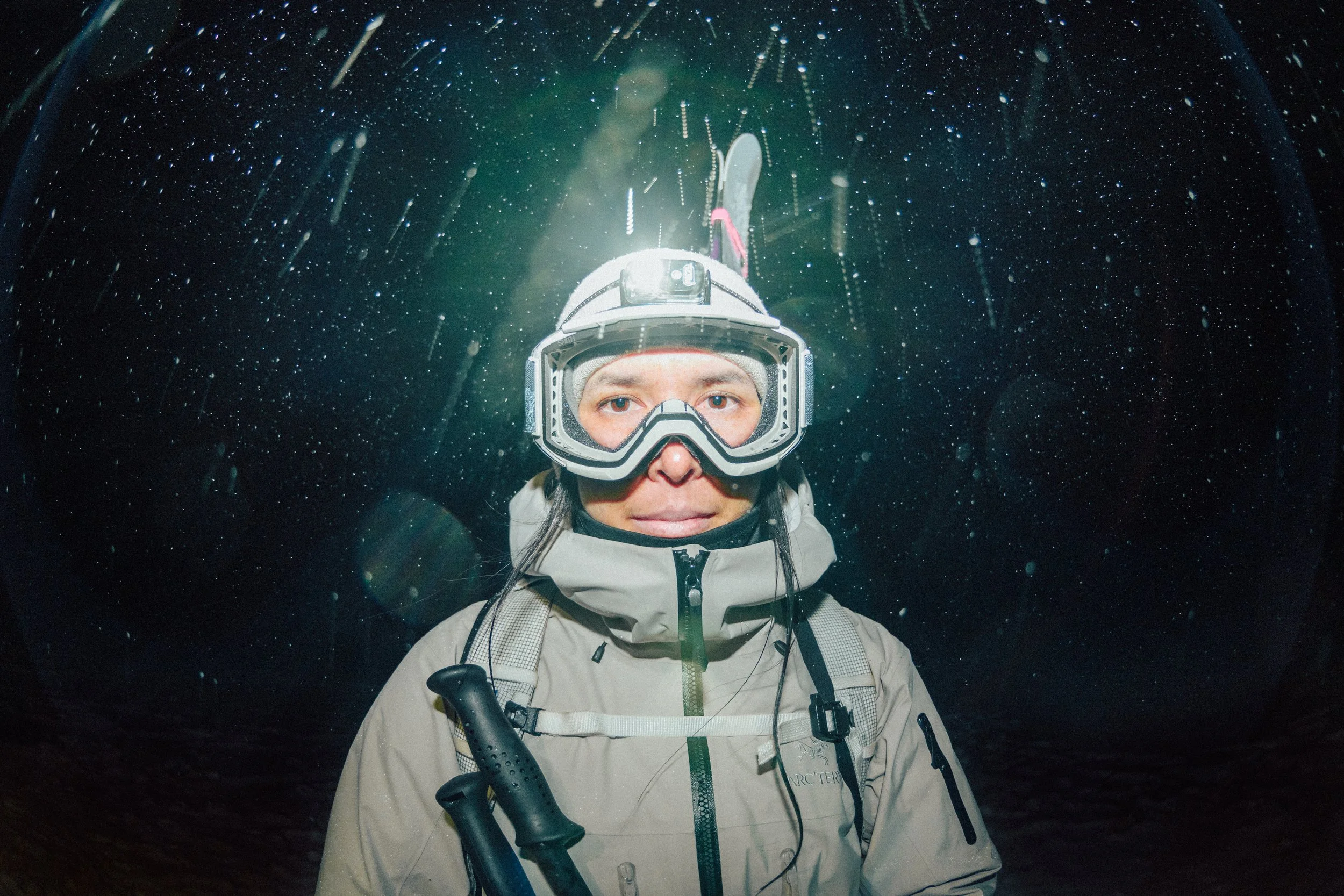 A person wearing a helmet and outdoor gear is standing in the rain at night, surrounded by falling rain and illuminated by a light source behind them.
