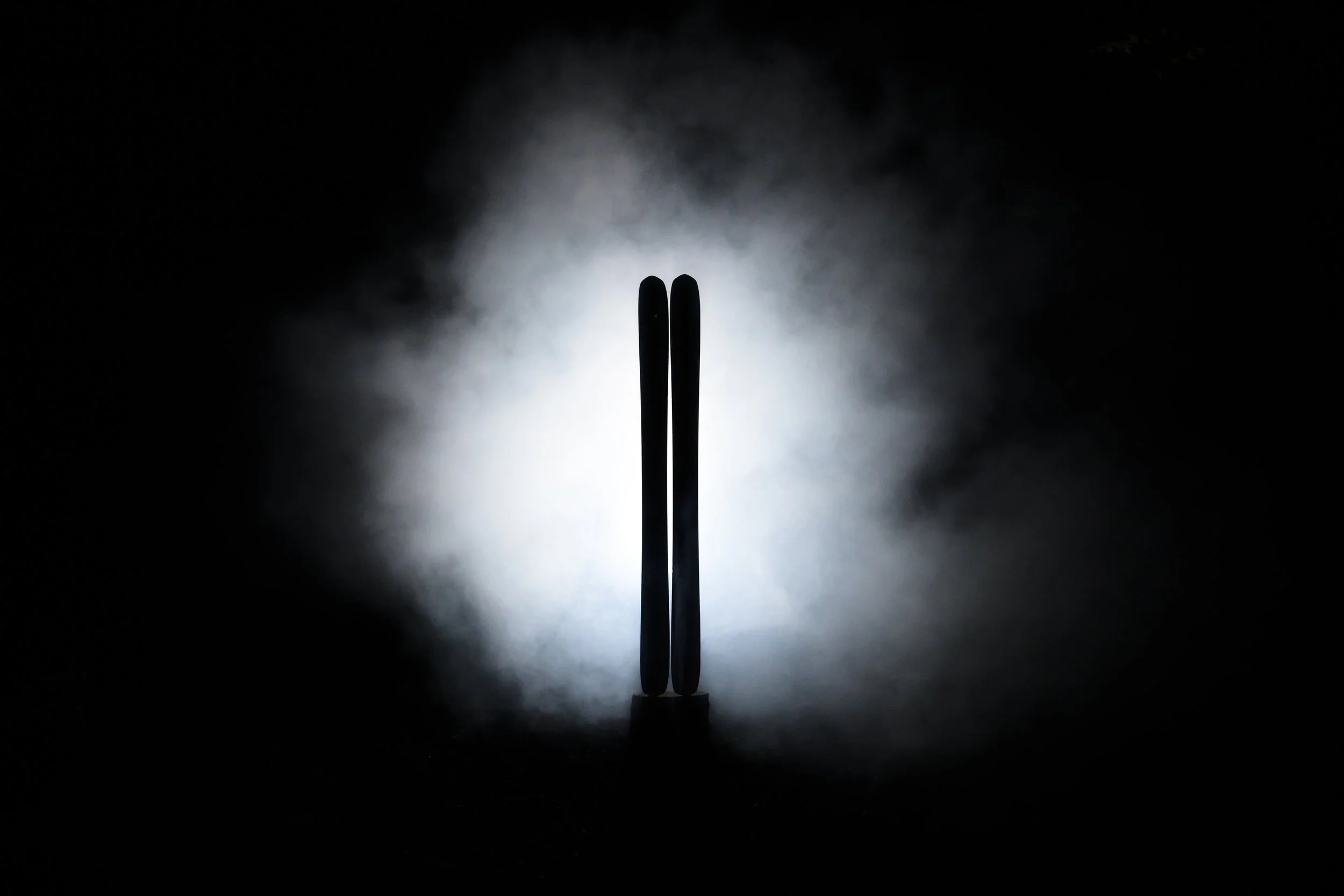 Two sticks or utensils standing vertically in front of a bright light with dark background, creating a silhouette and a glow around them.