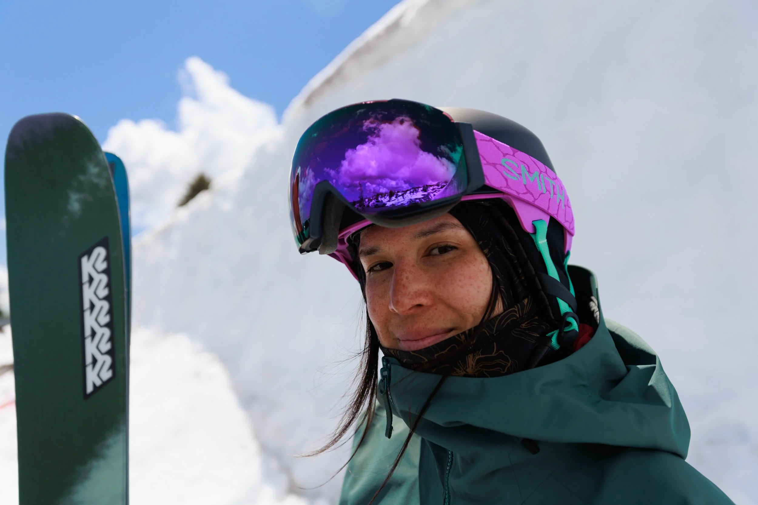 A woman wearing a pink ski helmet and mirrored ski goggles, smiling as she leans on her skis in a snowy mountain setting.