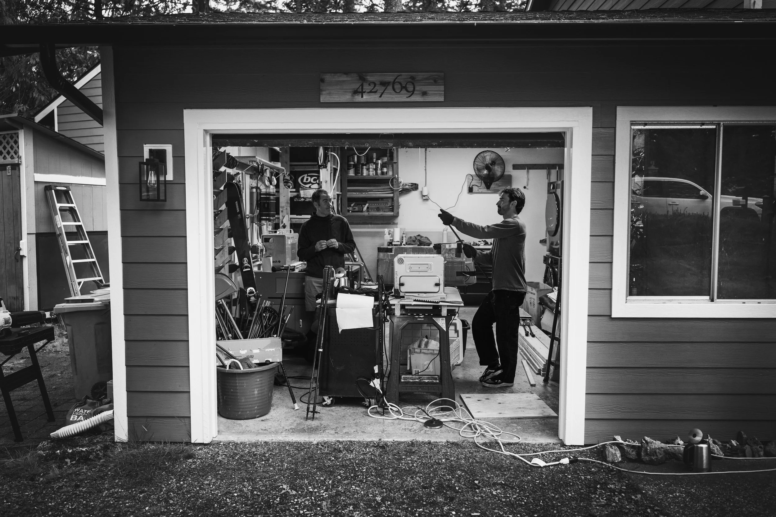 Black and white photo of two men in a garage, with one holding a long object and the other standing nearby, surrounded by tools and equipment, with an address sign 42769 above the garage door.