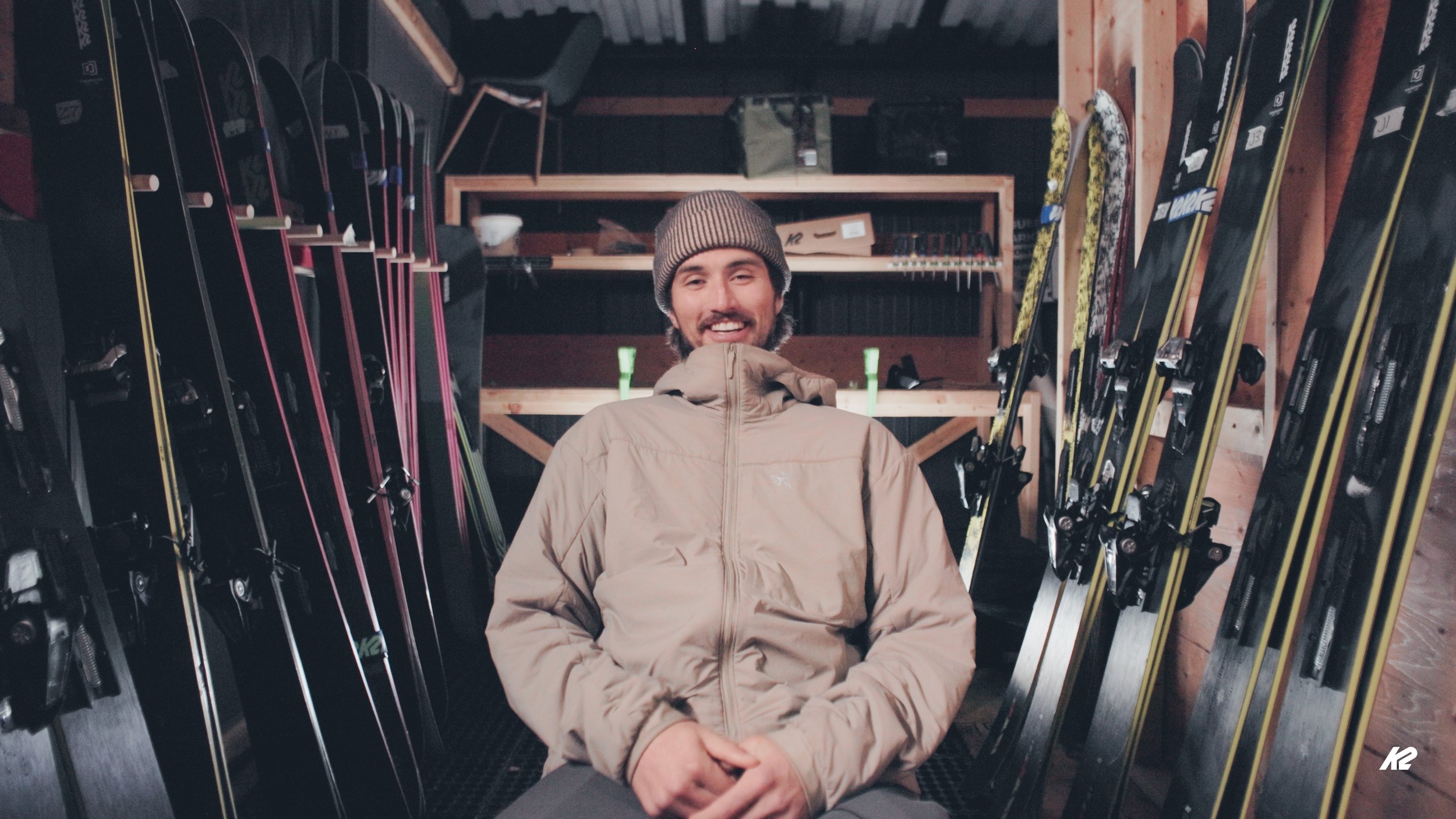A man in a tan jacket and brown knit hat sitting in a ski shop surrounded by skis on both sides, smiling at the camera.