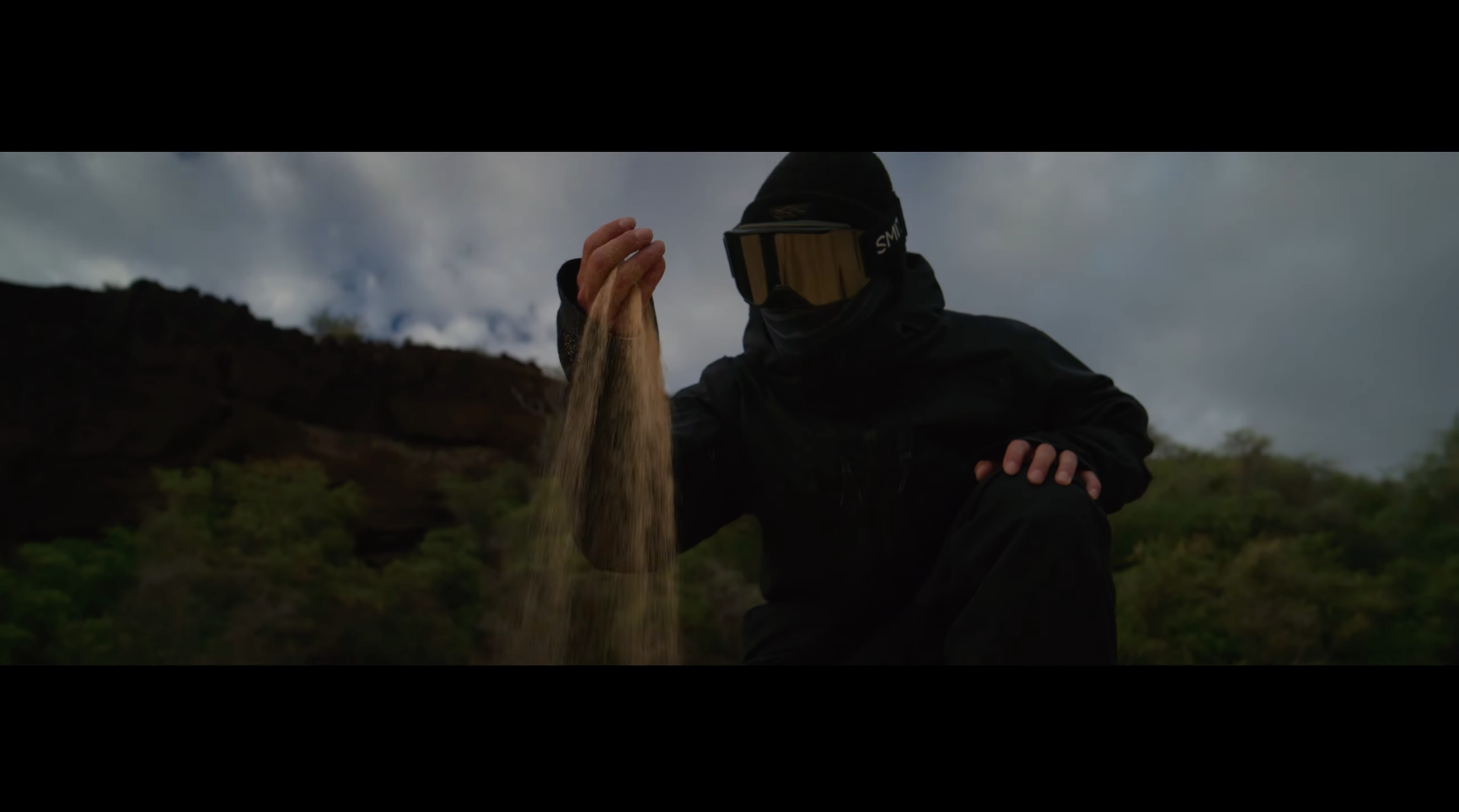 Person wearing black outdoor gear and goggles, squatting on a grassy hillside, pouring a stream of sand or dirt.