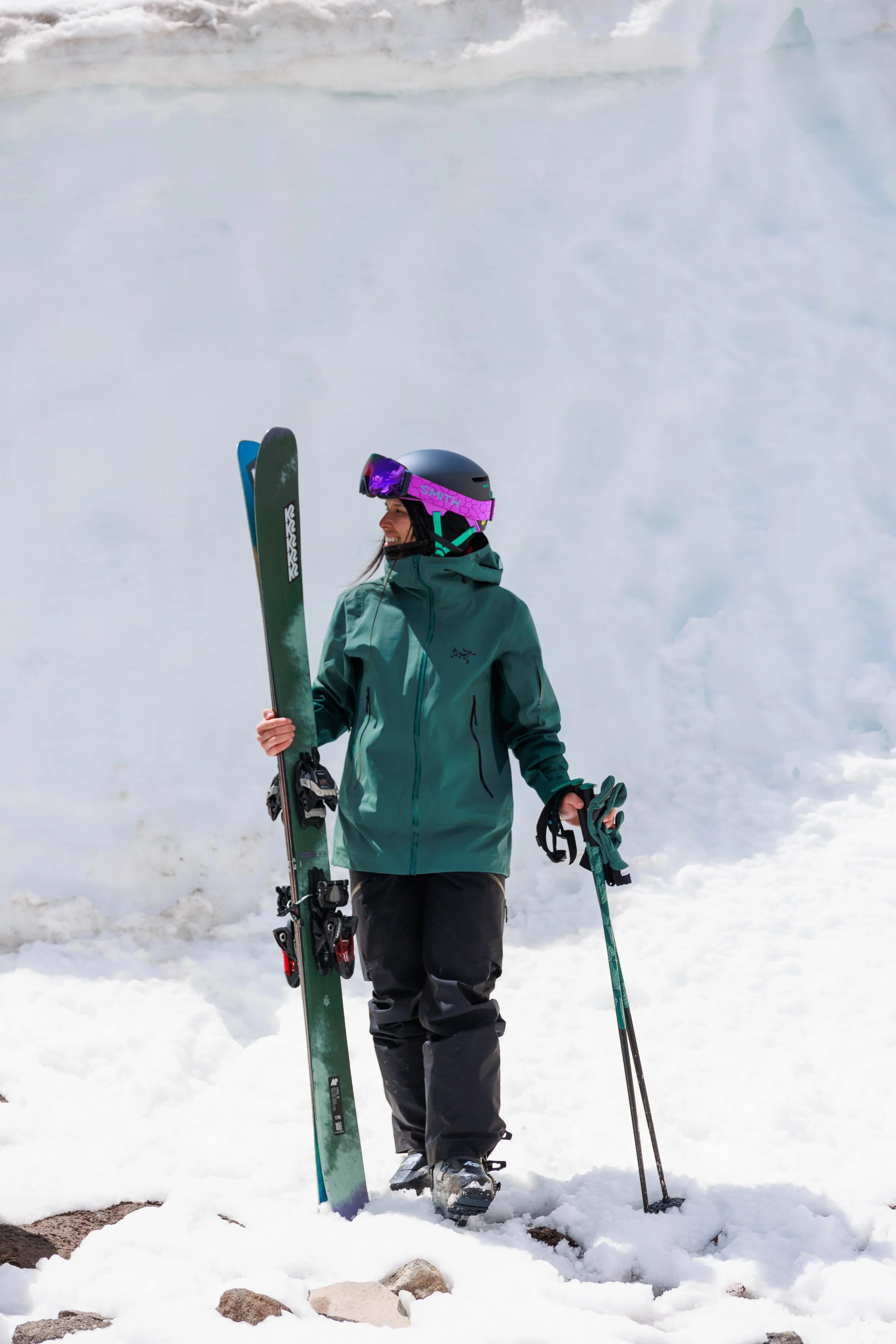 A person dressed in teal winter gear, wearing a black helmet with pink goggles, standing on snow with a pair of skis held upright in one hand and ski poles in the other, at a snowy mountain location.