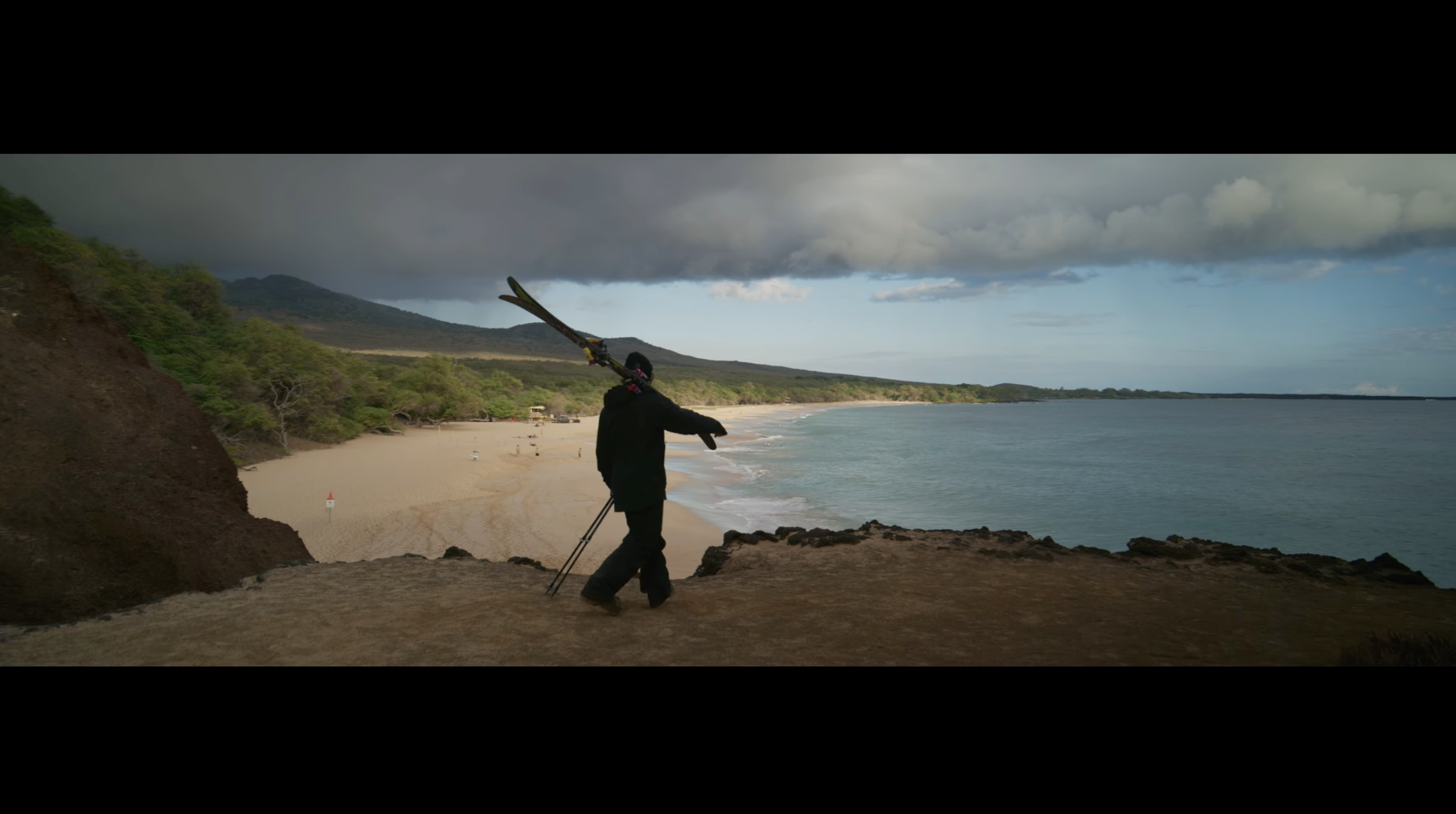 A person walking on a rocky ledge next to the beach, holding a long object over their shoulder, with a cloudy sky and distant mountains in the background.