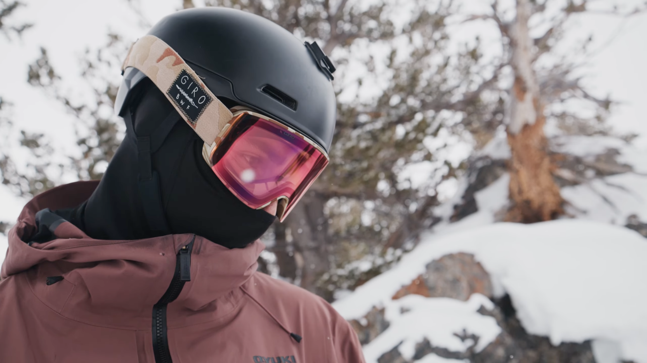 Person wearing a black helmet, pink goggles, black face covering, and a brown jacket in snowy mountain environment. Oyuki Jacket. A skier preparing to drop into a ski like.