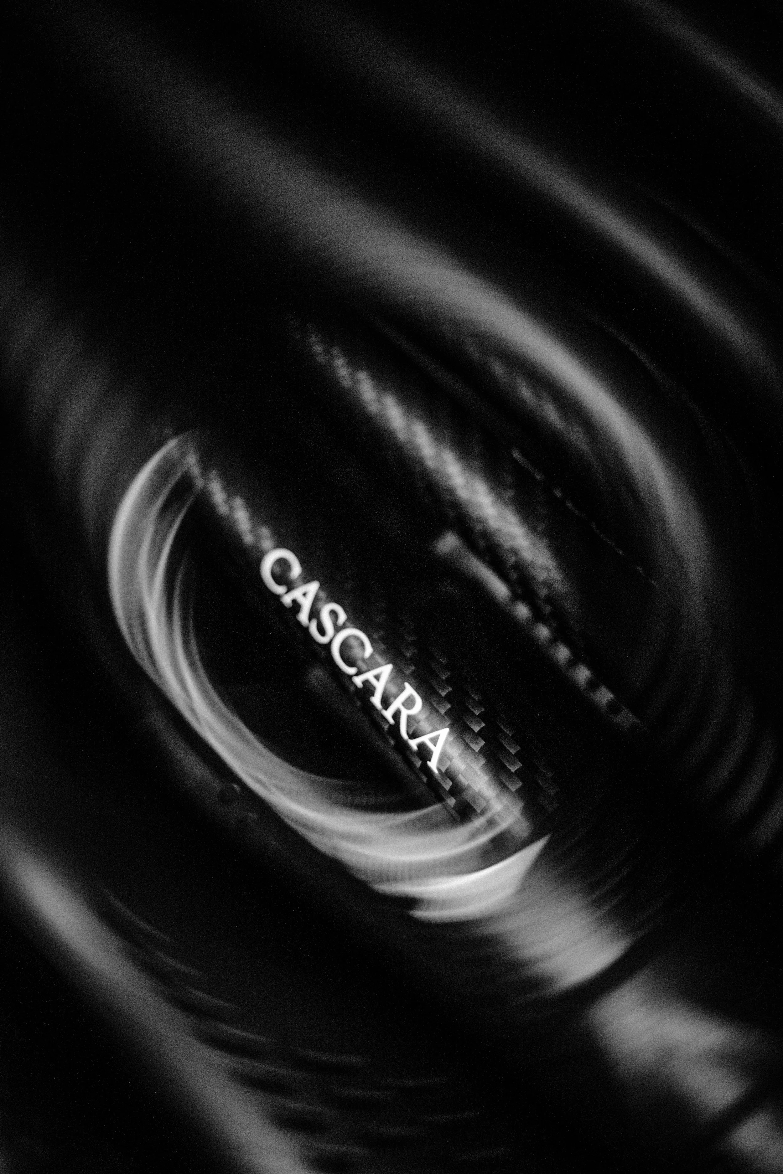 A close-up, black-and-white photograph of a glass bottle labeled 'CASCARILLA' viewed from the top, with a swirling pattern of light and shadow around it.
