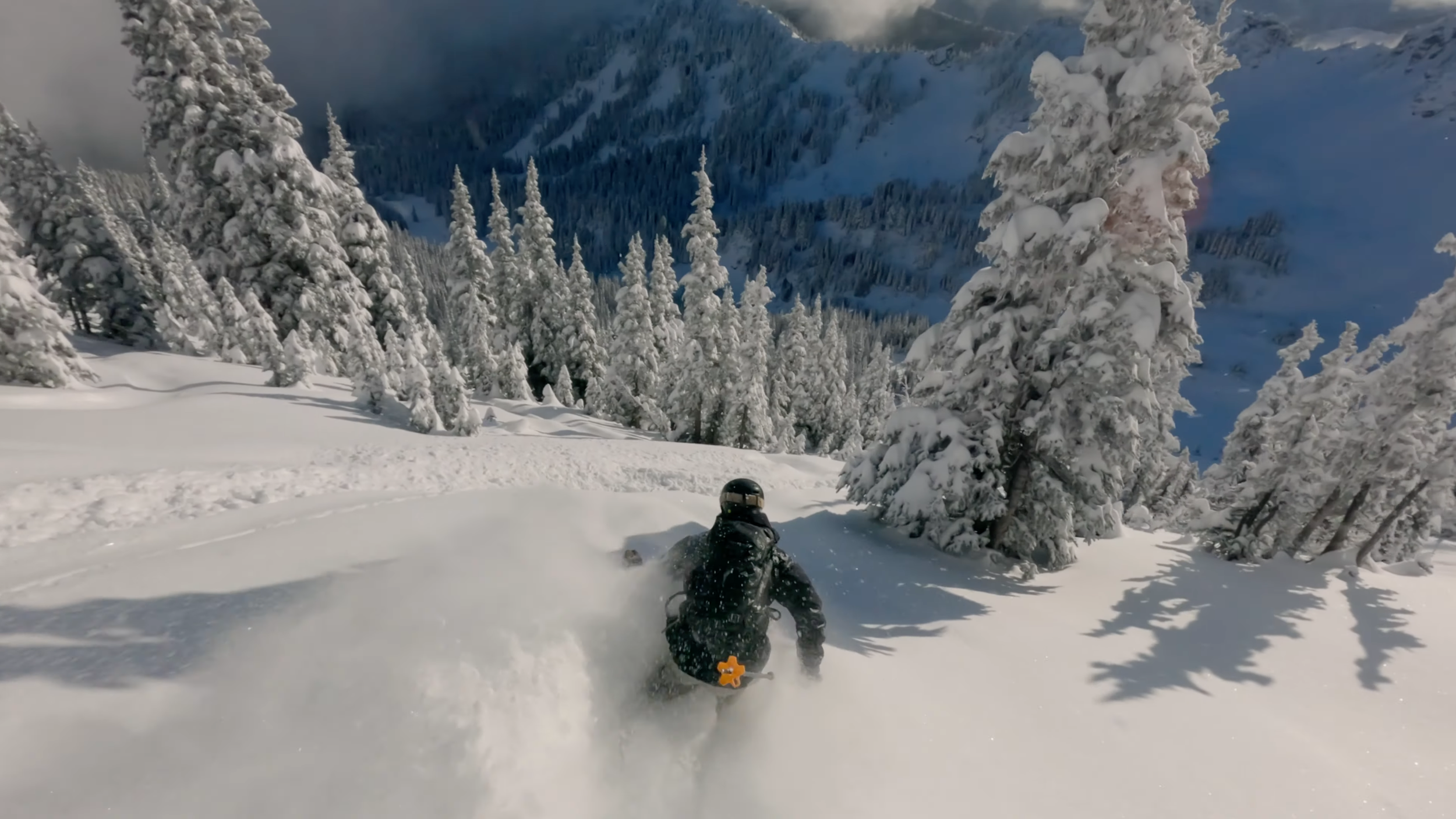 Person riding a snowboard down a snowy mountain surrounded by snow-covered trees under a cloudy sky. Deep powder turn.