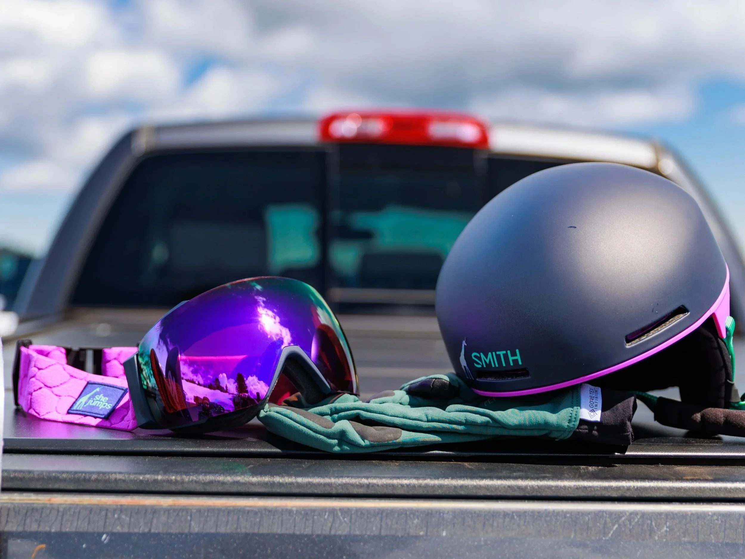 Helmets and goggles for snow sports placed on a black surface, with a vehicle and cloudy sky in the background.