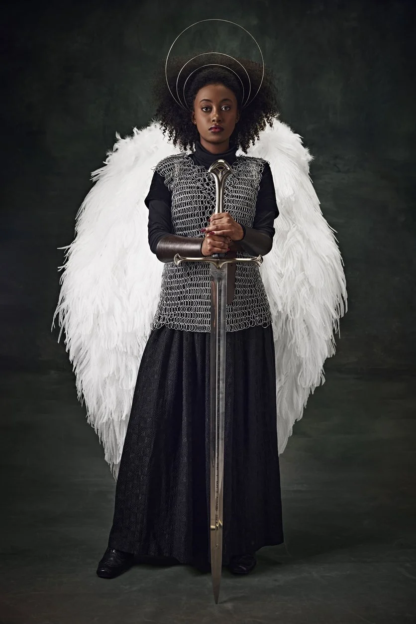 Black female warrior with white feathery wings, wearing a long black dress and chainmail, holding a sword and looking at the viewer with a serious expression