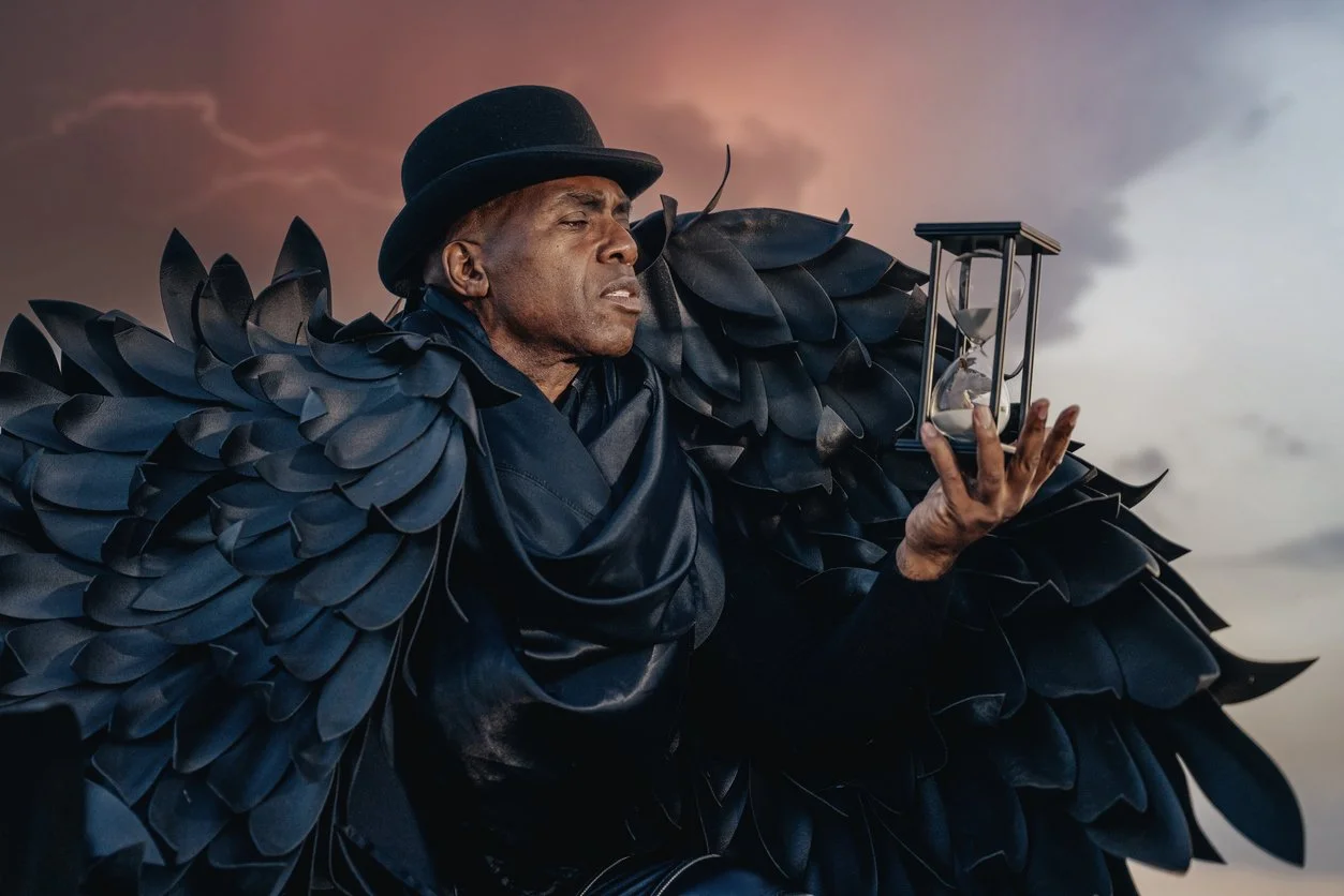 Middle-aged Black man with black feathery wings wearing a black bowler hat, holding an hourglass