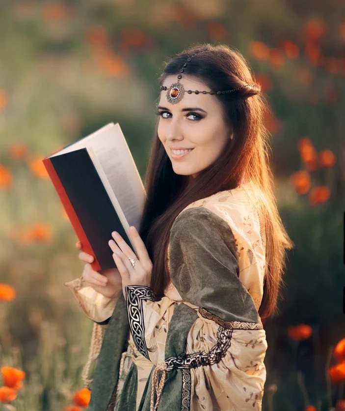 Pale brunette woman in a medieval dress reading a book in a meadow and looking flirtatiously at the viewer