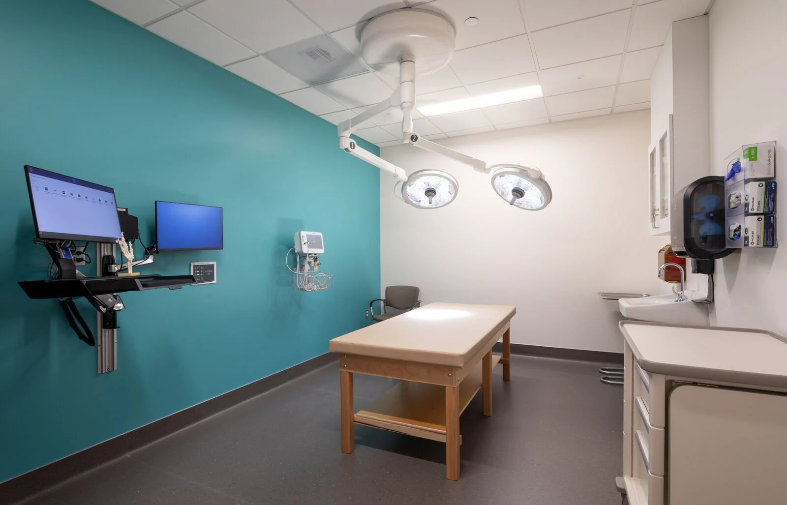 Empty hospital or clinic examination room with a medical bed, wall-mounted monitors, medical equipment, and surgical lights.