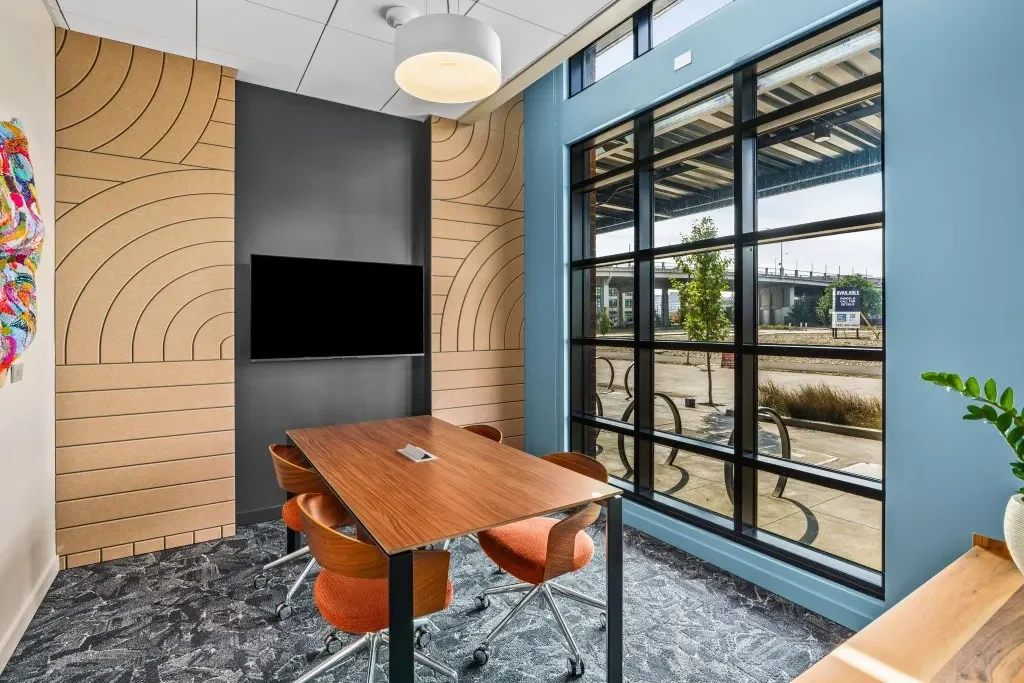 A modern conference room with a wooden table, four orange chairs with wheels, a flat-screen TV mounted on a dark gray wall, wooden wall panels with curved patterns, a large window showing an outdoor scene, and a ceiling light fixture.