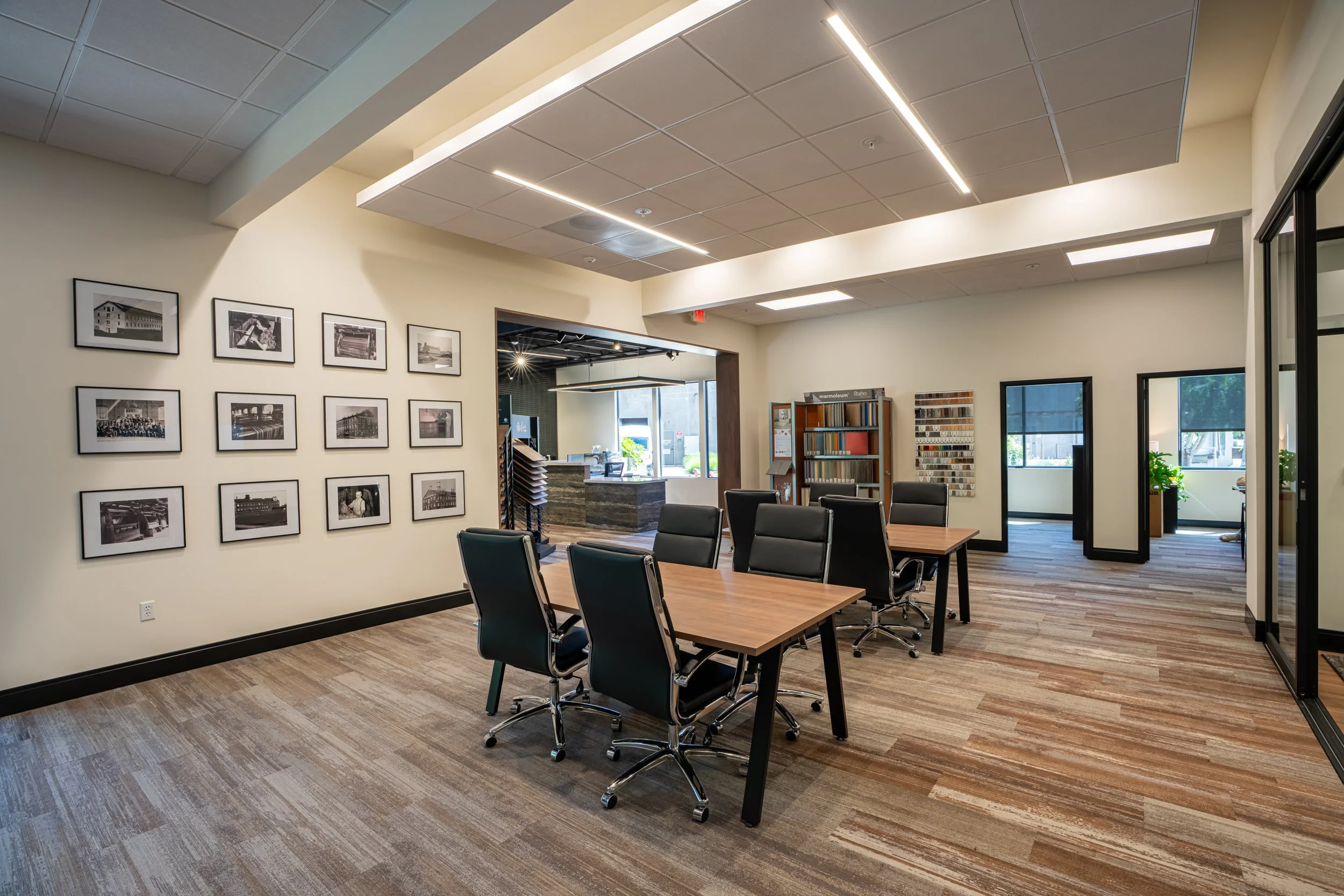 A modern conference room with black chairs around a wooden table, artwork on the wall, and a reception area. Large windows let in natural light, and the ceiling features built-in lighting.