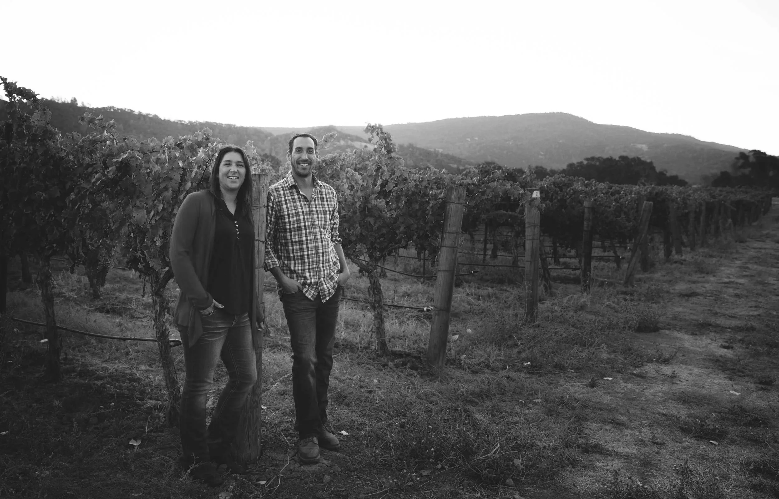 A black and white photo of a man and a woman standing in a vineyard with hills in the background, smiling at the camera.