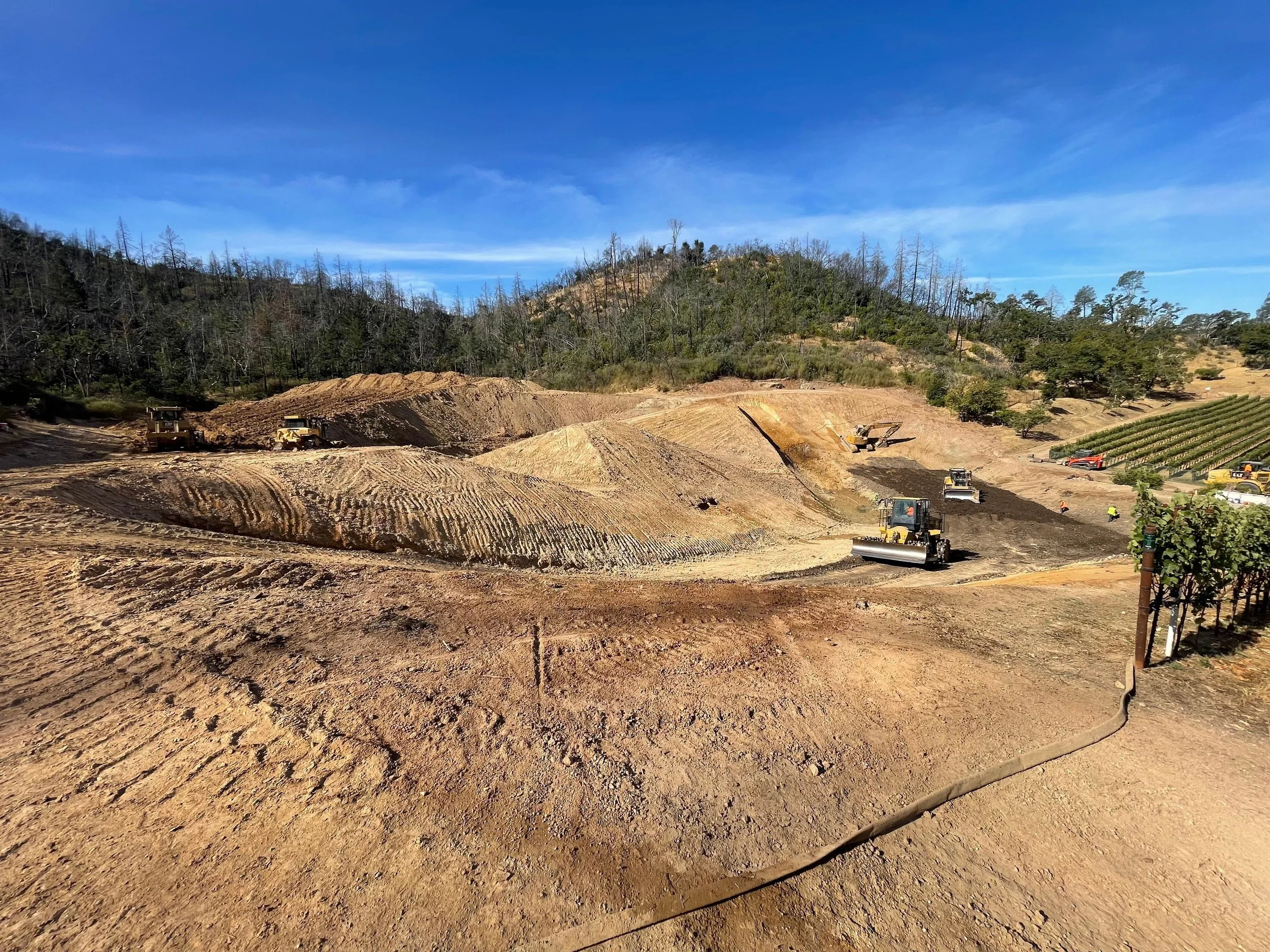 Construction site with bulldozers and earth-moving equipment, hillside with sparse trees, and a partly cloudy sky.
