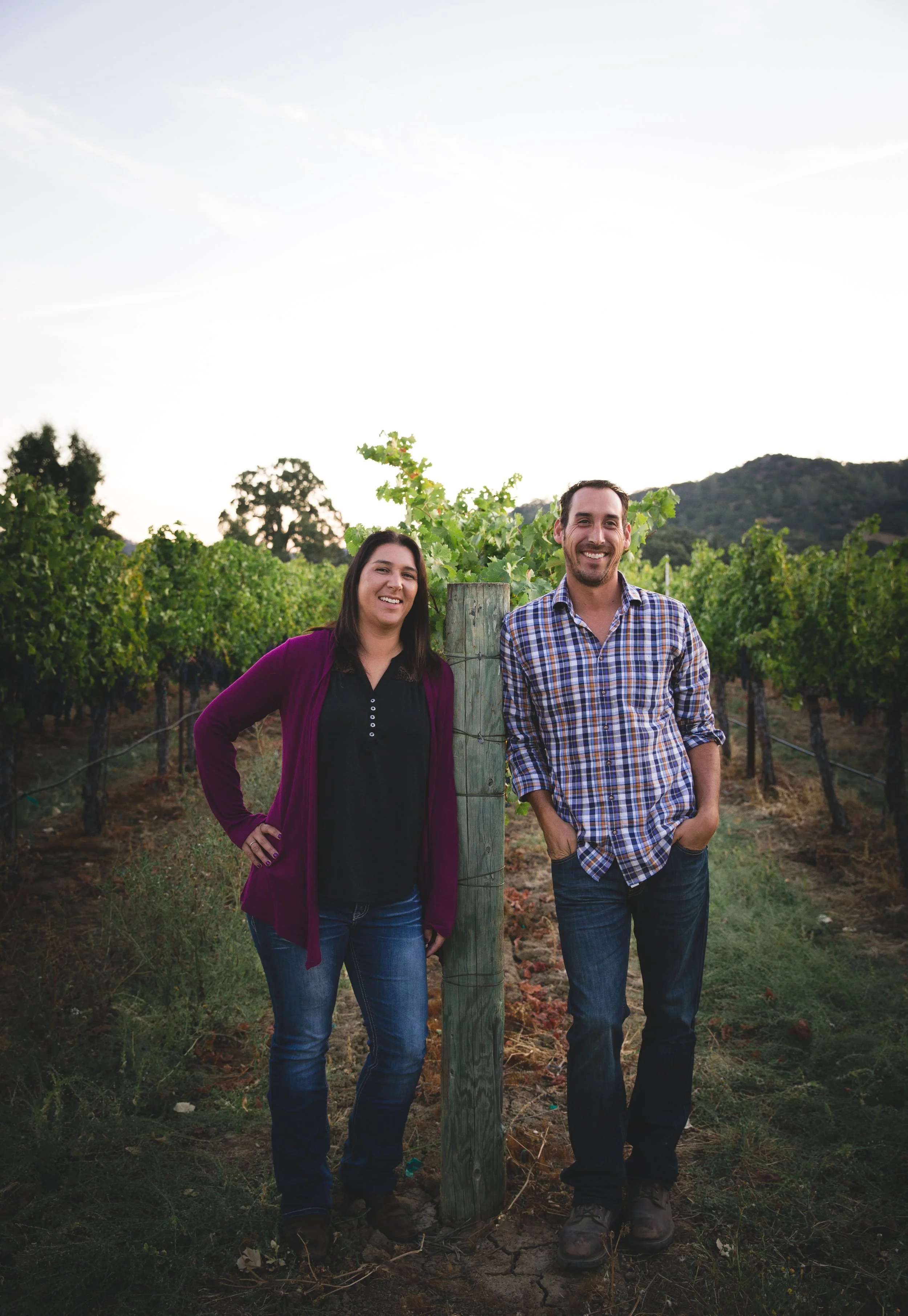 A man and woman standing in a vineyard outdoors, smiling, with grapevines and a mountain in the background, and a wooden post between them.