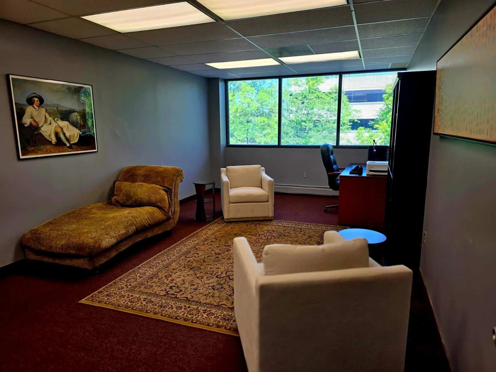 Office room with beige chairs, a chaise lounge, a desk with a black office chair, and large windows showing green trees outside.