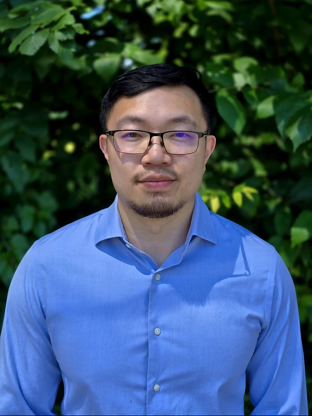 A man with short black hair, glasses, and a beard, wearing a blue button-up shirt, standing in front of green leafy plants.