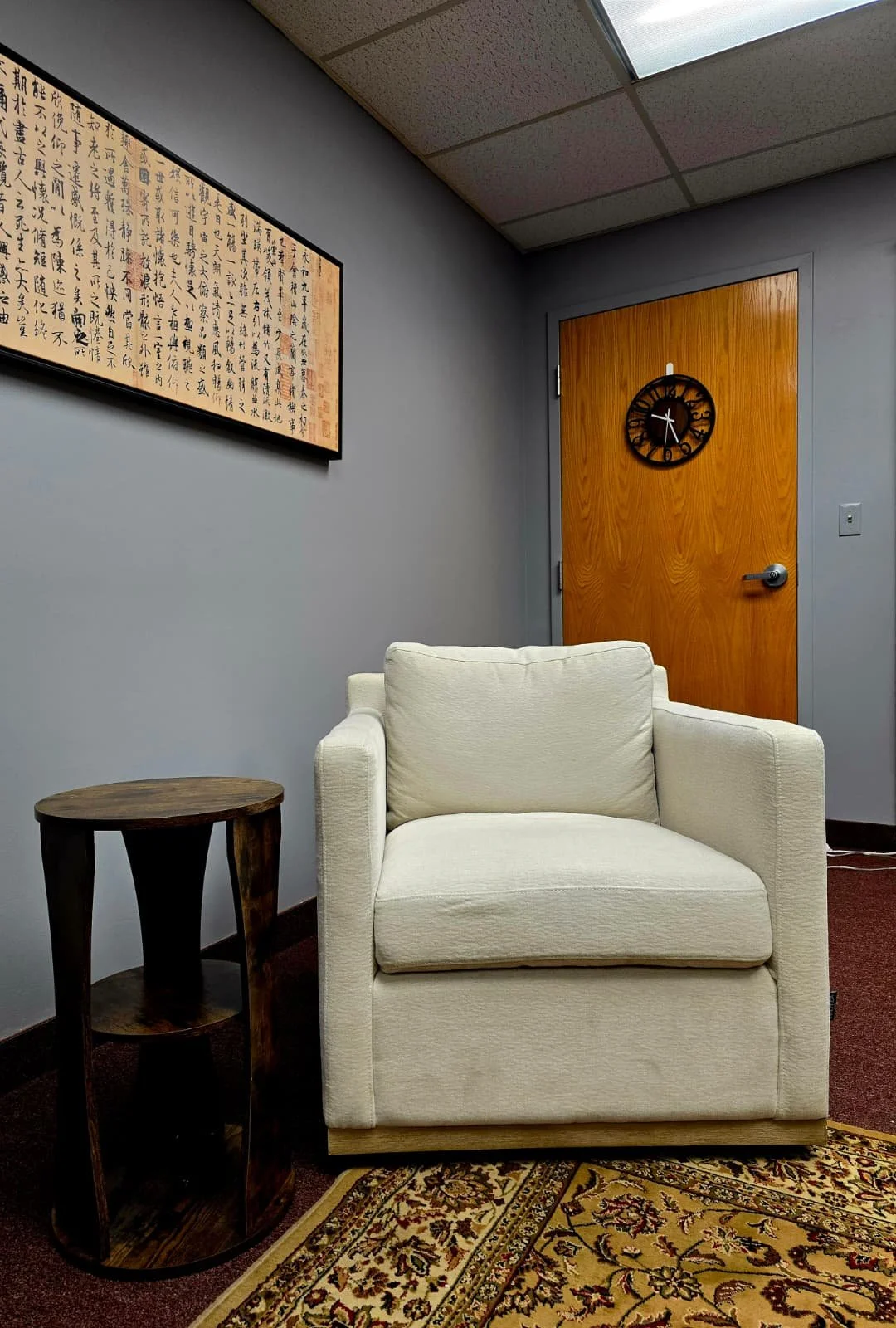A cozy room corner with a white armchair, a small wooden side table, a beige and brown patterned rug, a wooden door with a black clock, a gray wall, framed artistic writing, and a brown carpet.