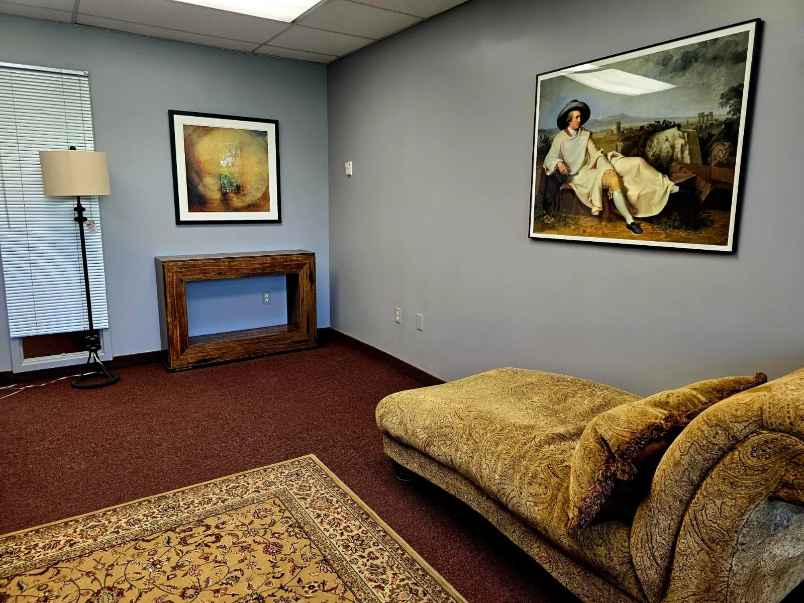 An interior room with a framed painting of a Goethe sitting outdoors on the wall, a wooden console table, a standing lamp, a window with blinds, a patterned area rug, and a patterned sofa with cushions.
