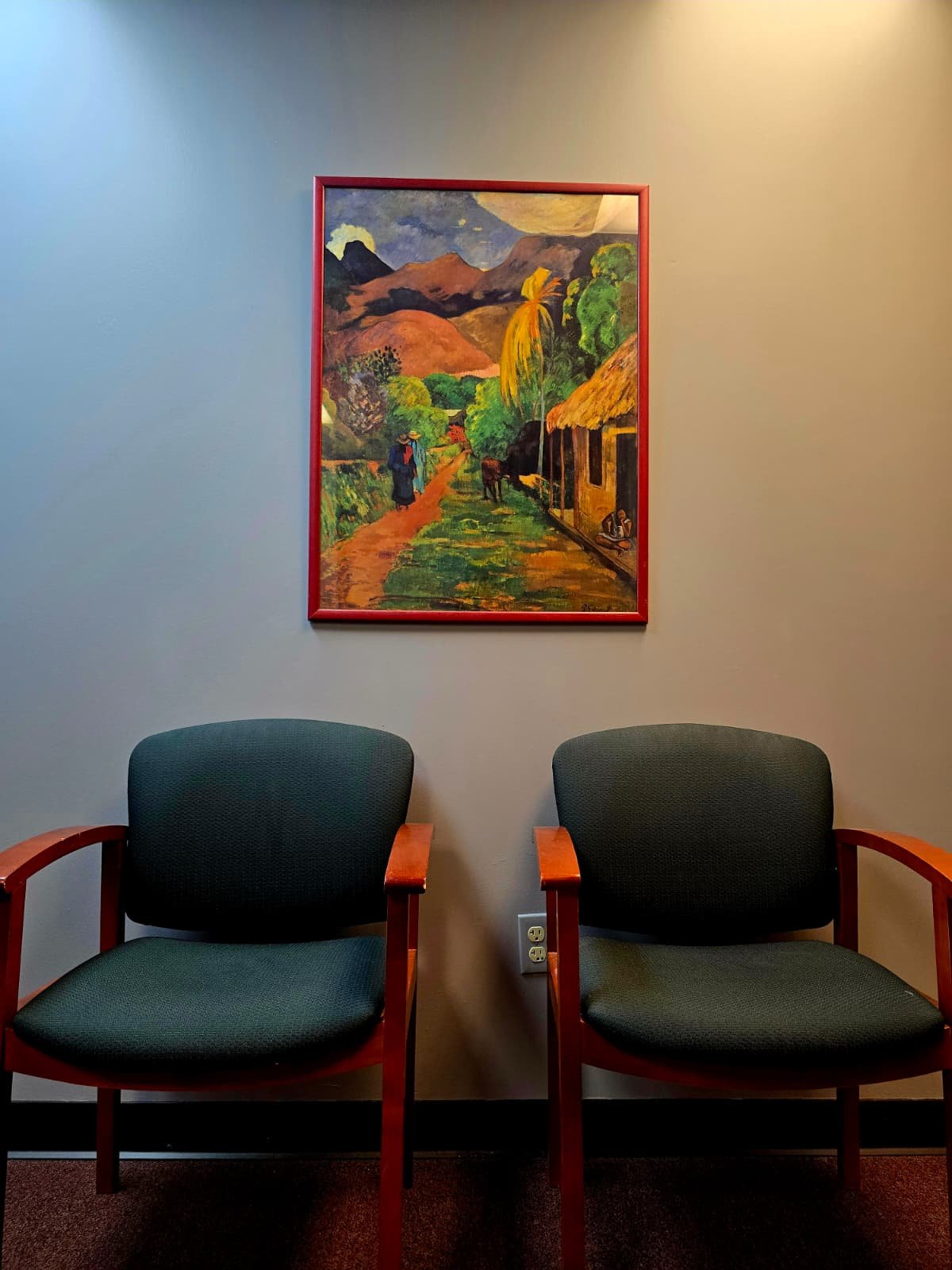 Two green upholstered chairs with wooden armrests are placed side by side in front of a wall with a framed colorful landscape painting. The painting depicts a rural scene with mountains, trees, a thatched house, and a person walking along a dirt path.