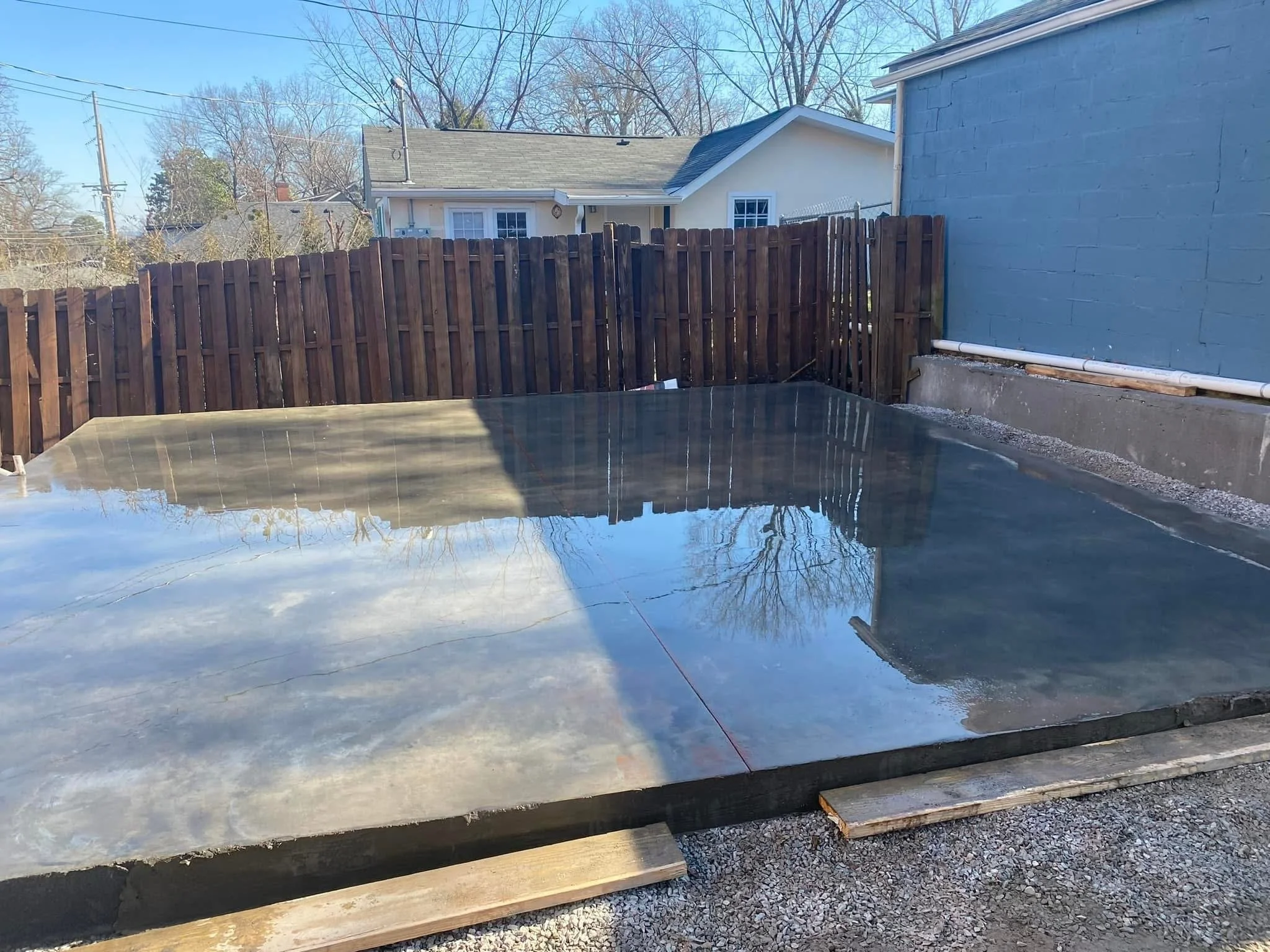 A backyard with a wooden fence, a concrete patio, and a house in the background. The patio is wet, reflecting the blue sky. There are leafless trees and utility poles visible beyond the fence.