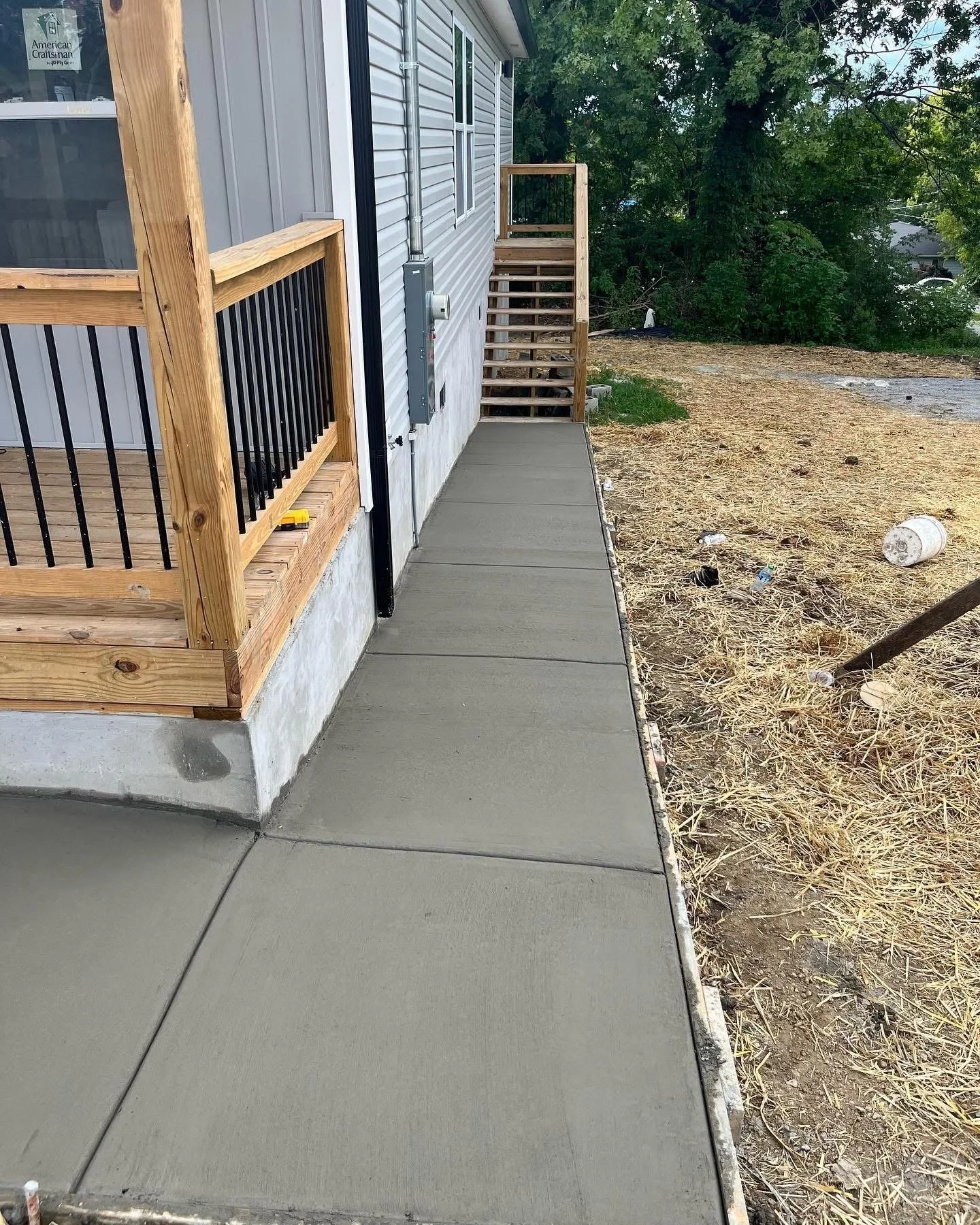 Newly poured concrete sidewalk next to a house with a partially built wooden staircase leading to a second level. The house has gray siding and window with a sticker that says 'American Craftsman.' There is a view of a yard with dry grass, a large tr