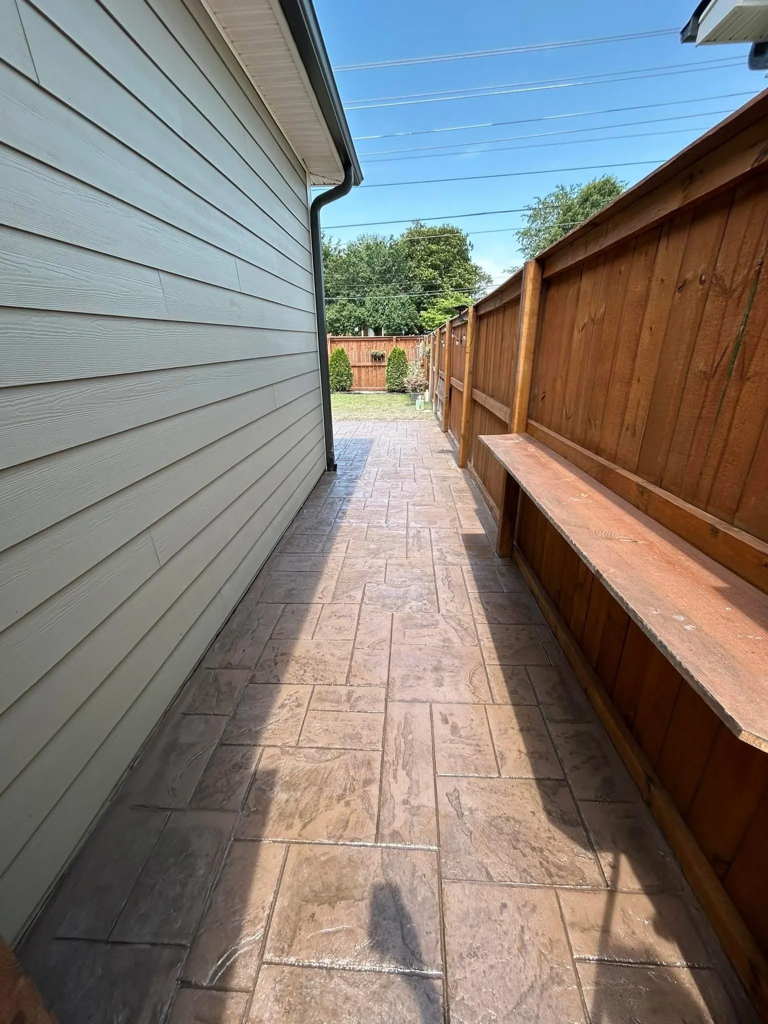 A narrow outdoor walkway with stamped concrete tiles on the ground, bordered by the side of a house with beige vinyl siding on the left and a wooden fence on the right. The sky is clear and blue with visible power lines.