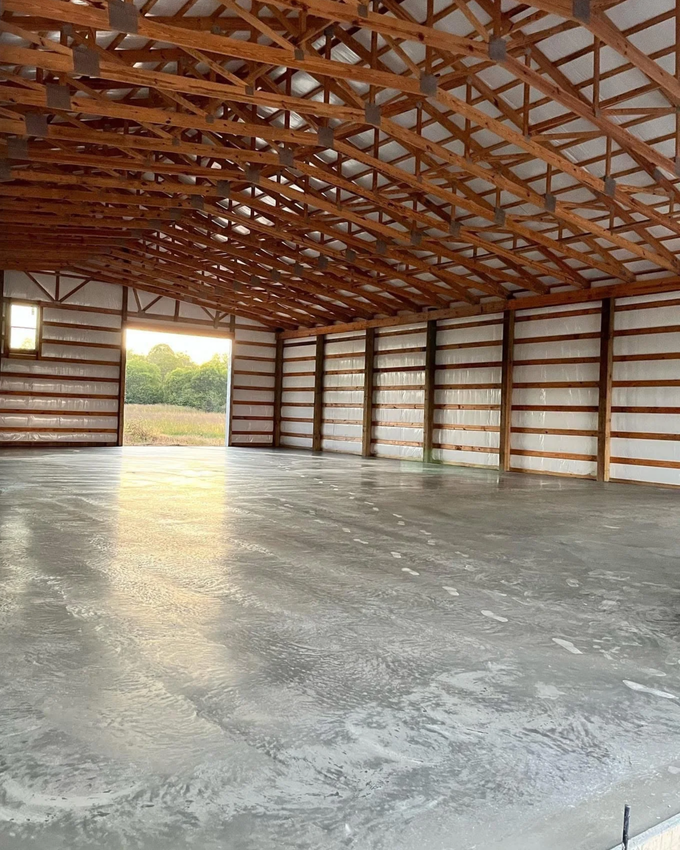 Interior of an unfinished barn with a concrete floor, wooden ceiling trusses, and walls made of metal siding, with open large door revealing a green field outside.