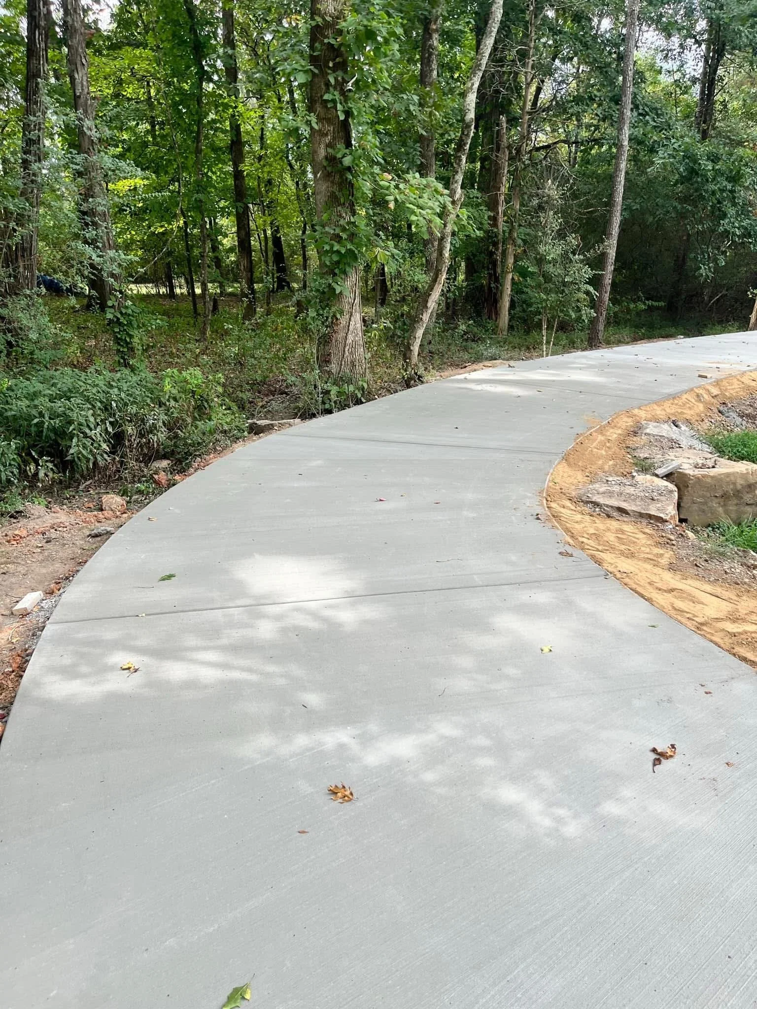Newly paved concrete pathway curving through a wooded area with trees and green foliage on both sides, and scattered leaves on the path.