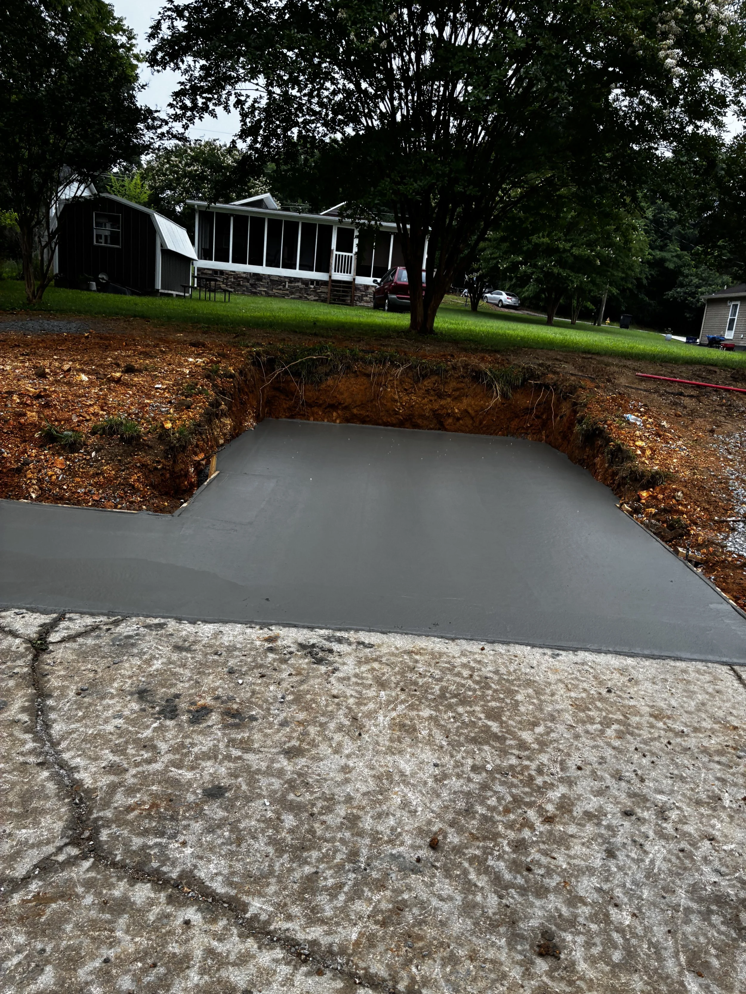 A freshly poured concrete slab on a sidewalk, with exposed dirt and construction tools nearby, in a suburban backyard with grass, trees, and houses in the background.