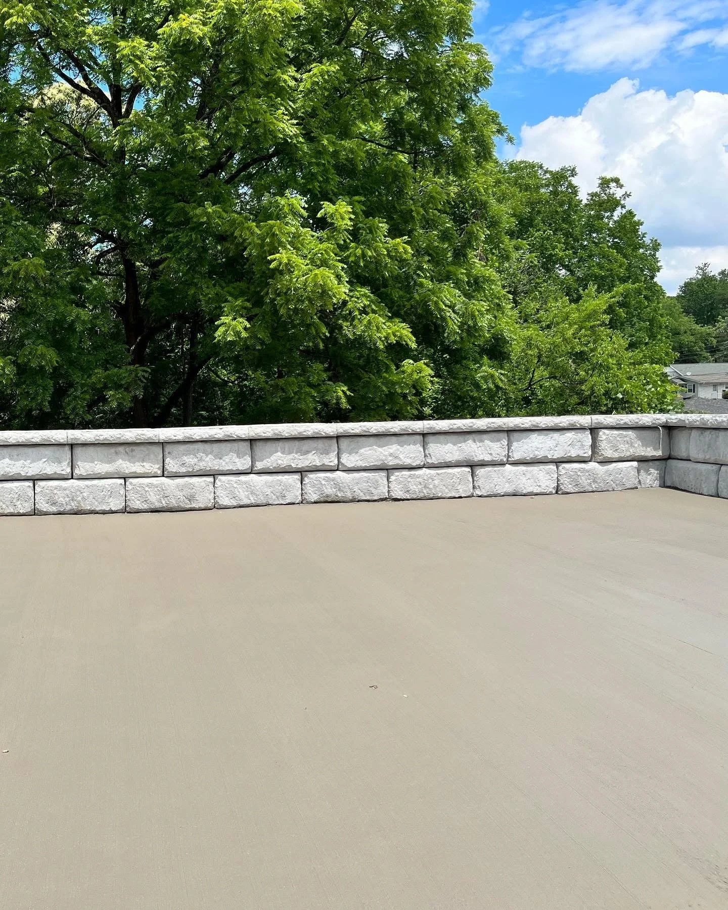 View of a concrete patio with a short stone wall and a background of green trees under a partly cloudy sky.