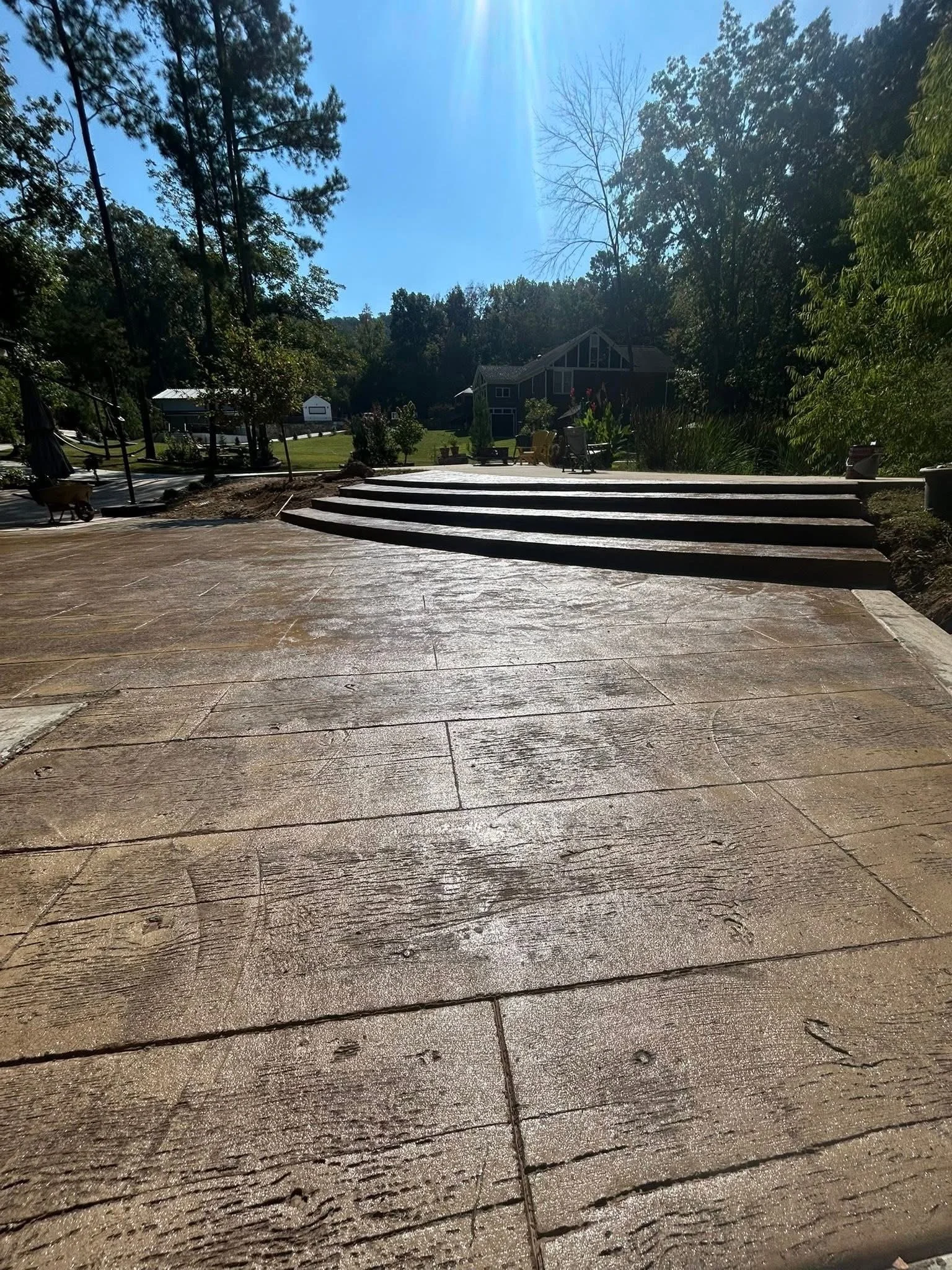 Image of a tiled backyard patio with stairs leading up to a grassy area with trees, bushes, and a house in the background on a sunny day.