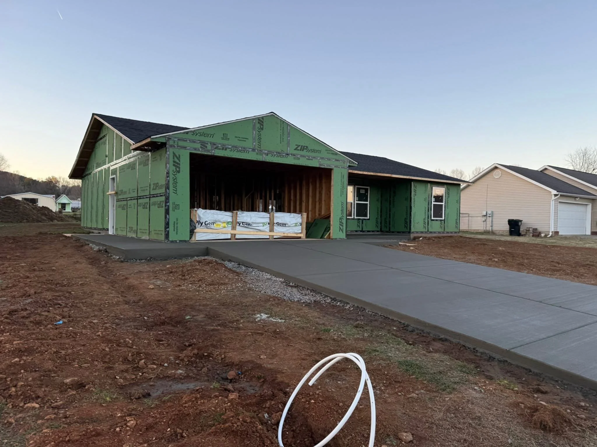 A house under construction with green exterior sheathing, a driveway, and neighboring houses in the background. The construction site has dirt and white pipes on the ground.