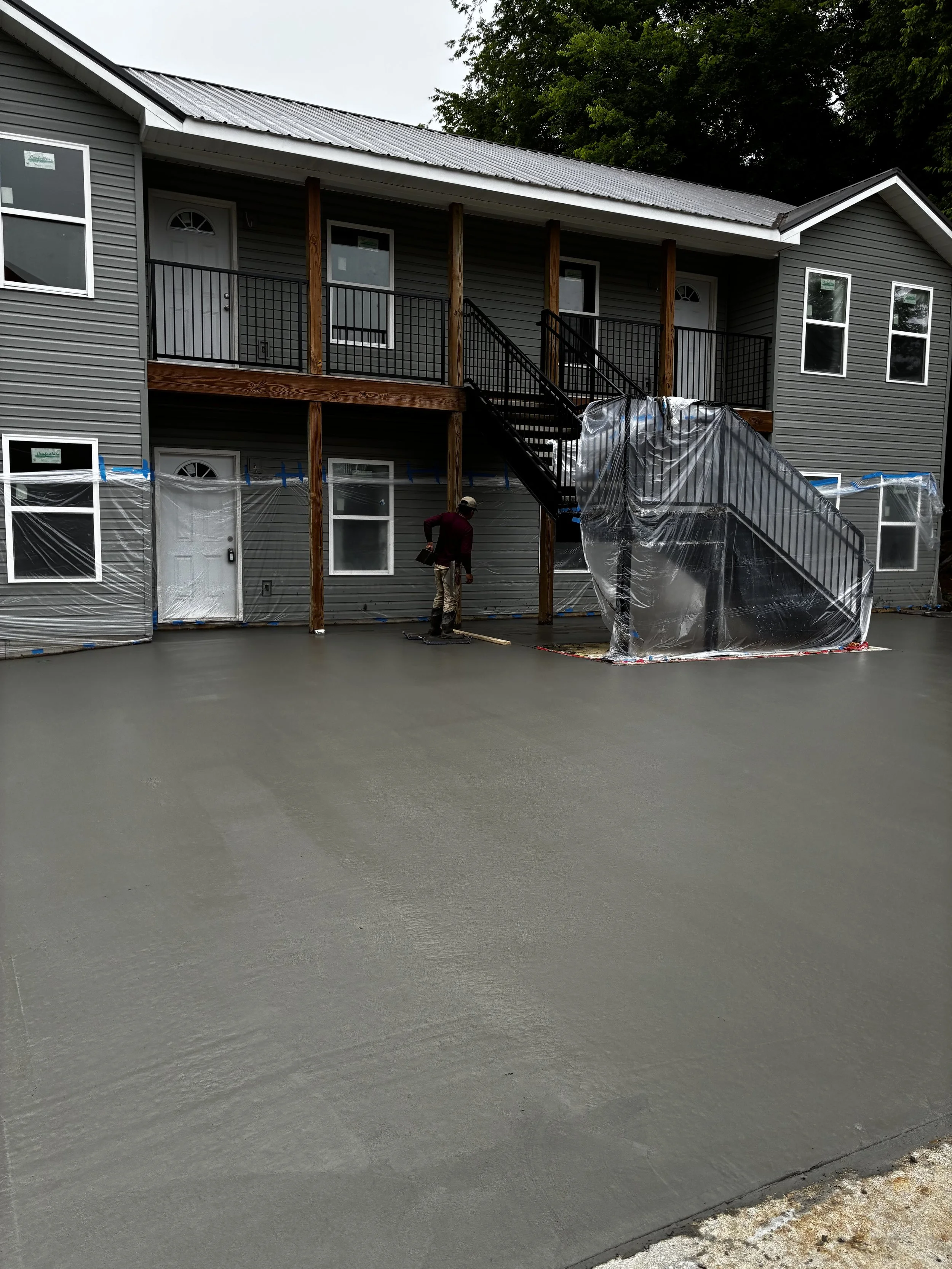 Newly poured gray concrete driveway in front of a gray two-story house with a deck and stairs under construction, protected by plastic sheeting, with a worker standing nearby.
