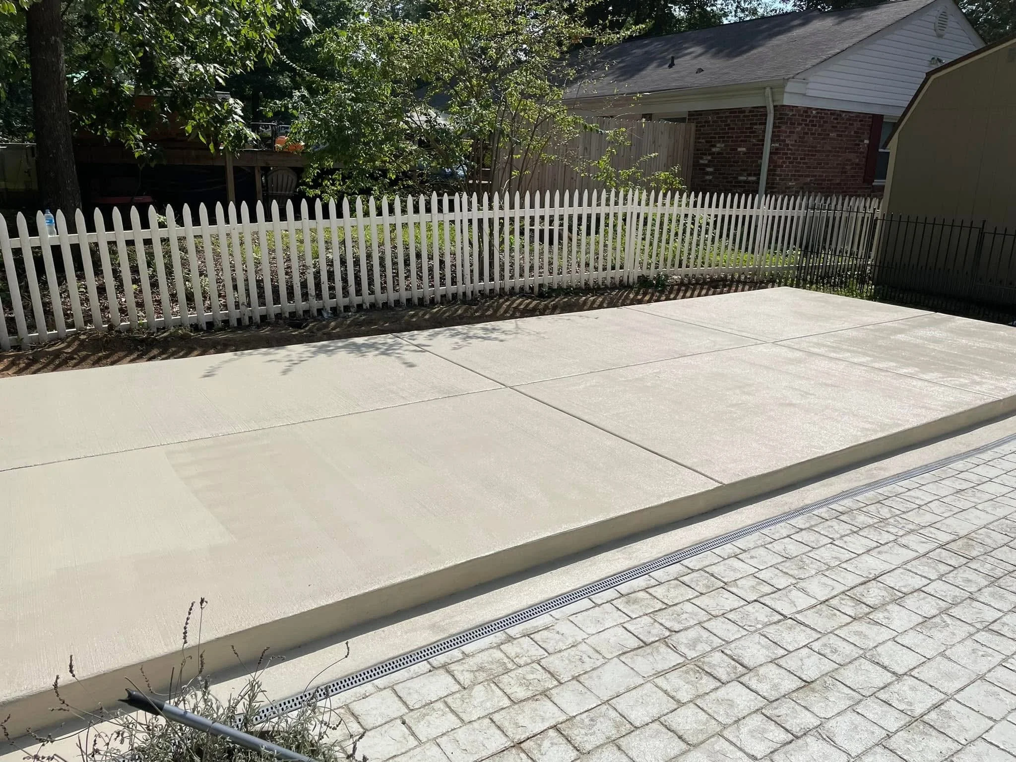 Backyard with a white picket fence, brick house, and green trees, concrete patio, and a drain grate in the ground.