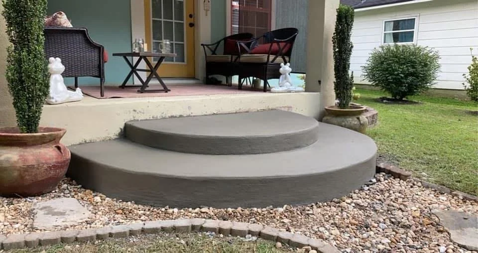 Concrete steps leading up to a porch, with potted plants and outdoor furniture on the porch and grassy yard in the background.