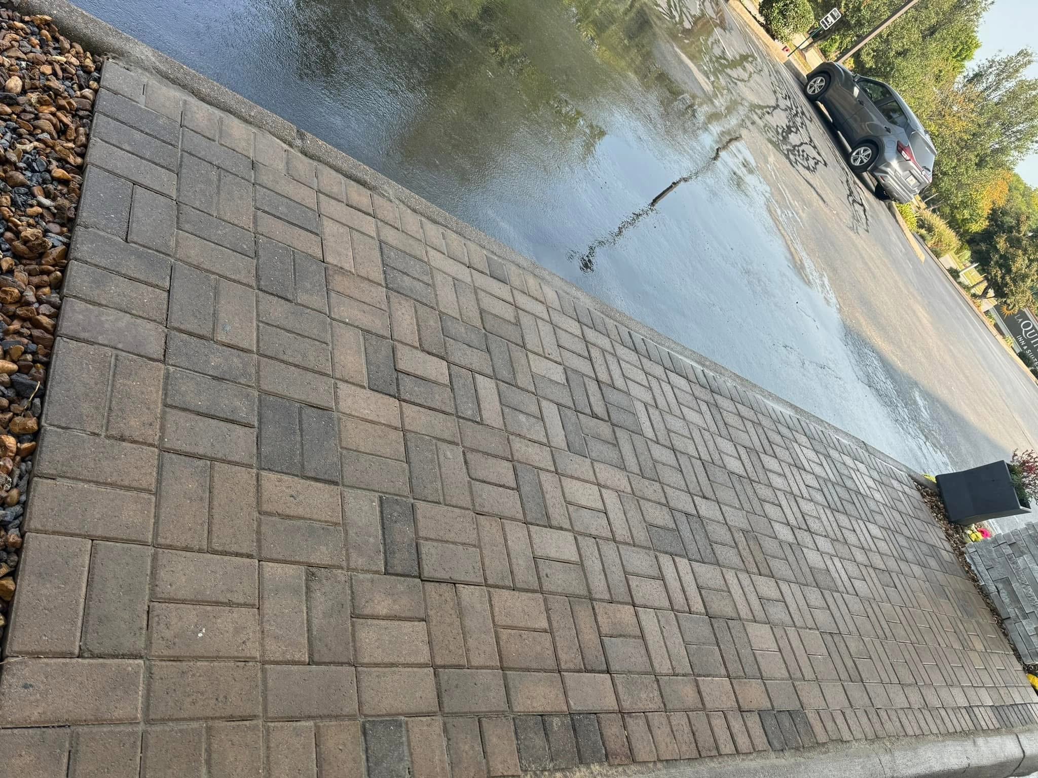 A parking lot with a wet surface, a marked sidewalk with bricks, and a silver SUV parked. Reflections of trees and cars are visible on the wet asphalt, with some trees and greenery in the background.