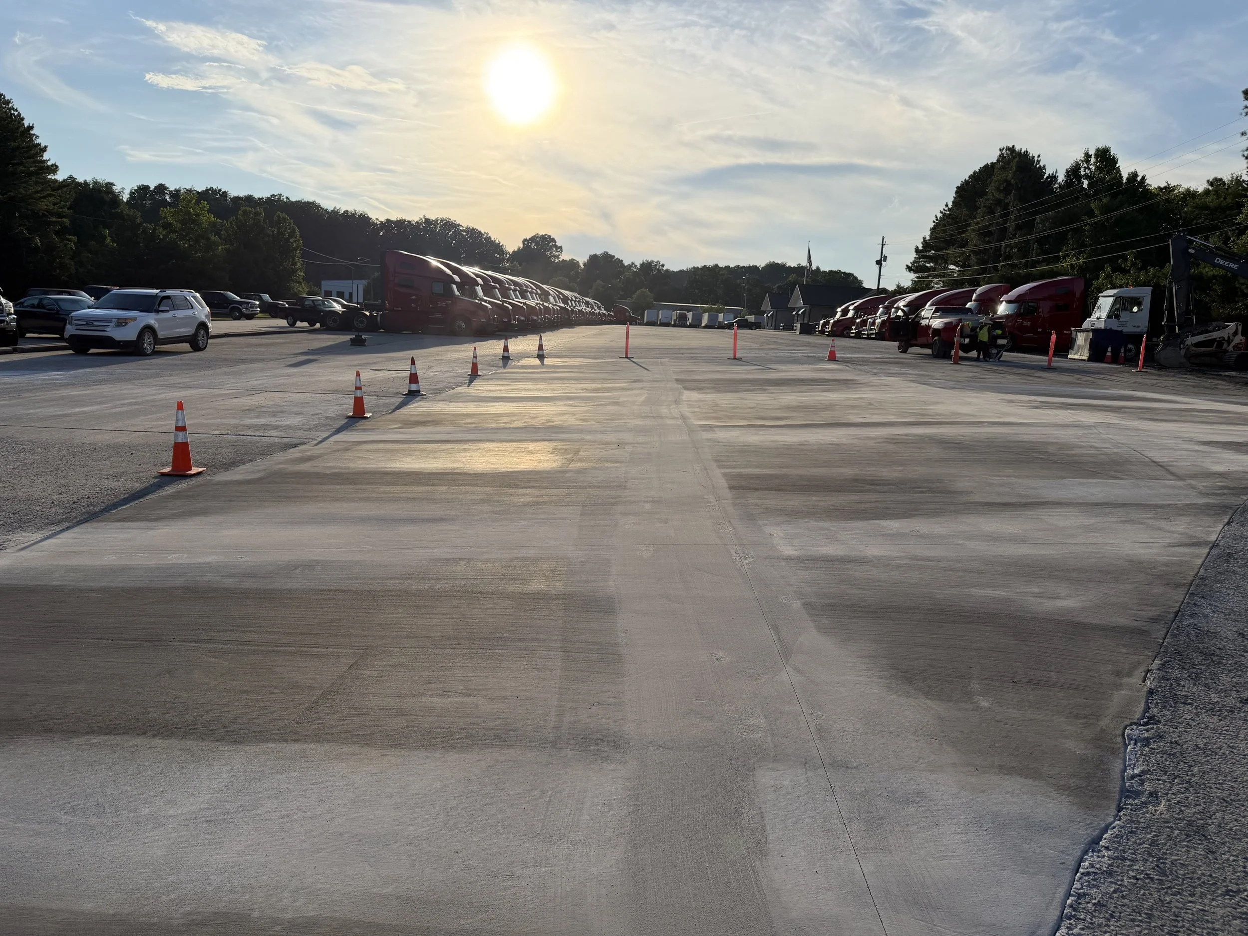 A large parking lot with several parked vehicles, including semi-trucks with red trailers and a white SUV, during sunset under a partly cloudy sky. People are seen near the vehicles, and orange traffic cones are placed across the lot.