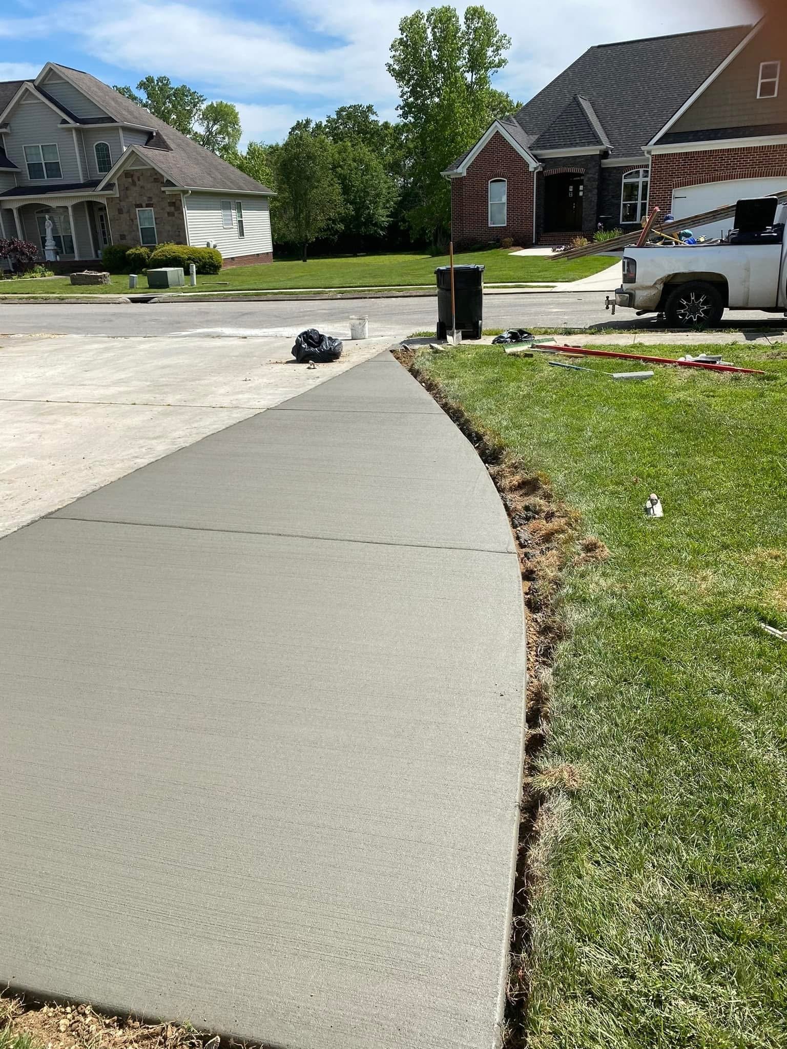 New concrete sidewalk next to grass in front yard of suburban neighborhood, with houses and trees in the background.