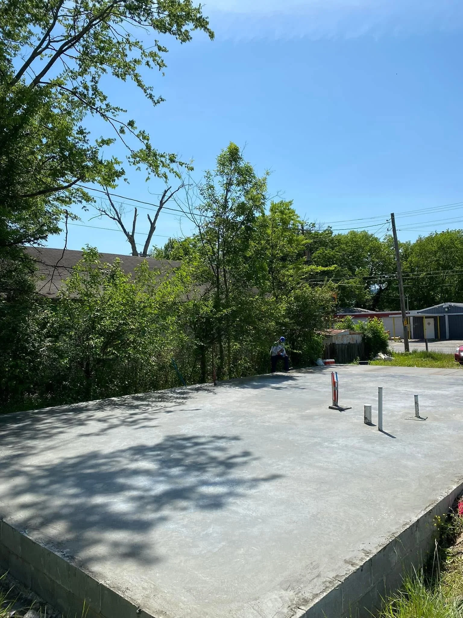A freshly poured concrete slab for a building foundation in a residential area, with safety posts and shadows from nearby trees.