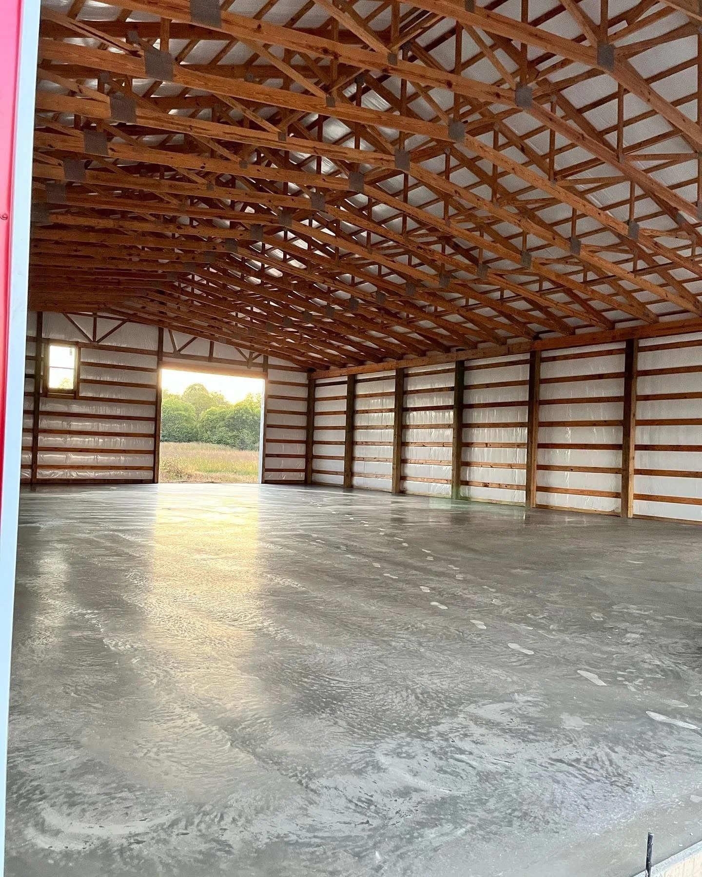 Interior of an unfinished barn with a concrete floor, wooden ceiling trusses, and walls made of metal siding, with open large door revealing a green field outside.