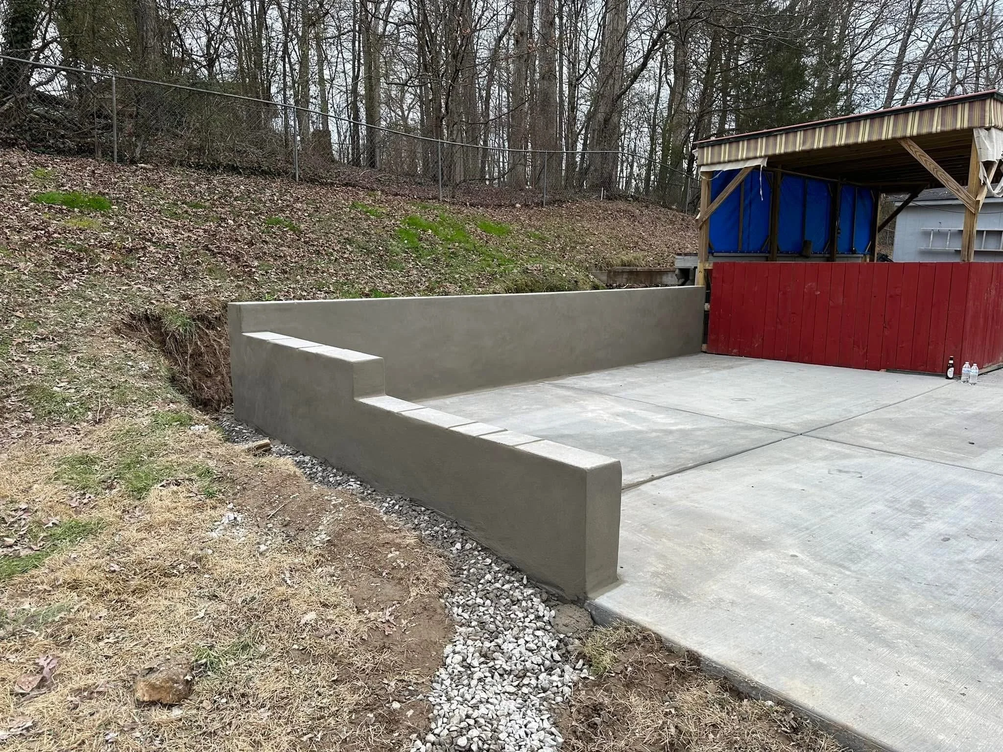 A concrete retaining wall and a concrete patio near a wooded hillside with leafless trees, a chain-link fence, and a small structure with a canopy and a red wooden fence.